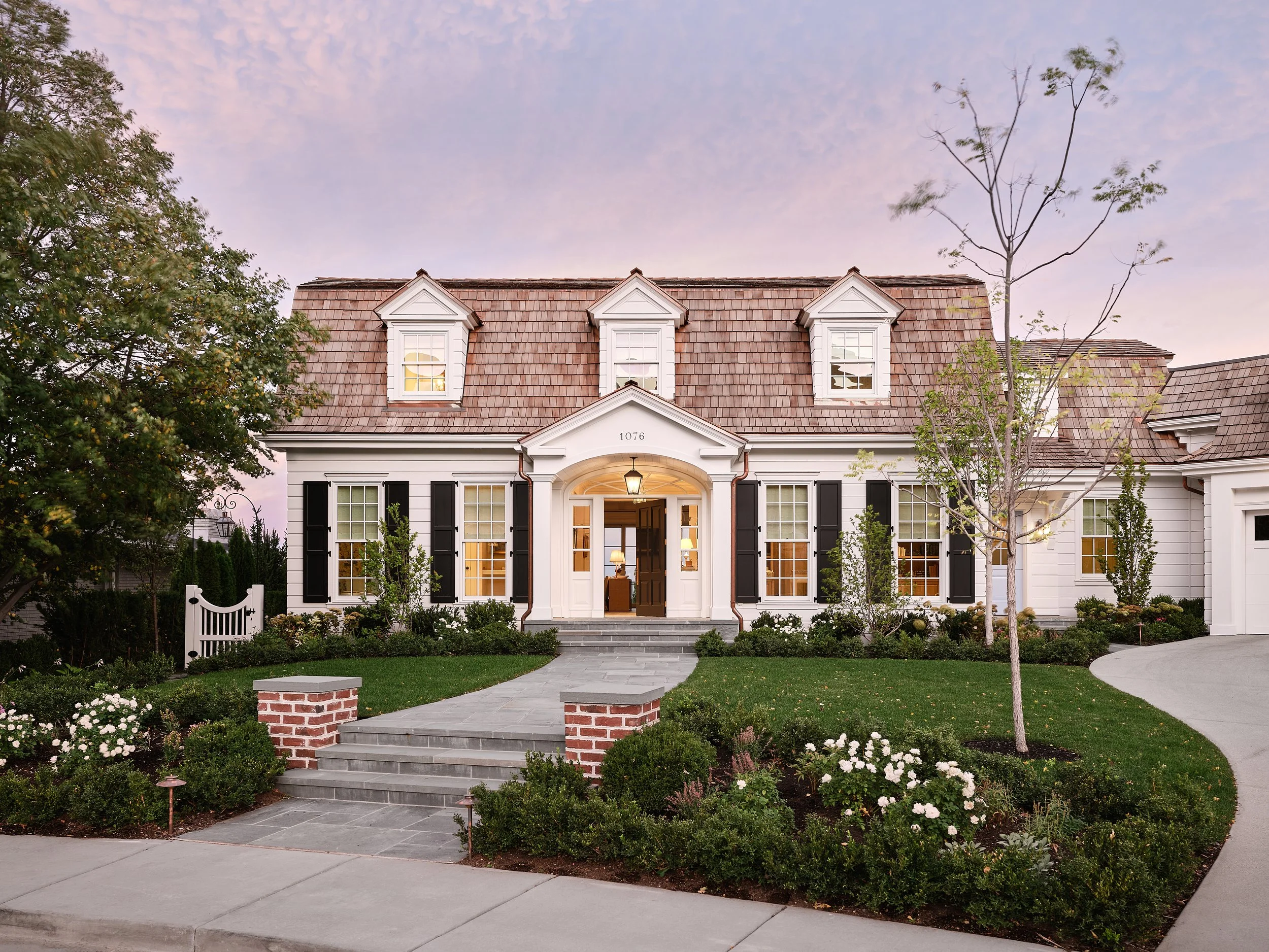 Front view of a white house with black shutters, brown shingled roof, and a manicured front yard with trees and flowers, during sunset.