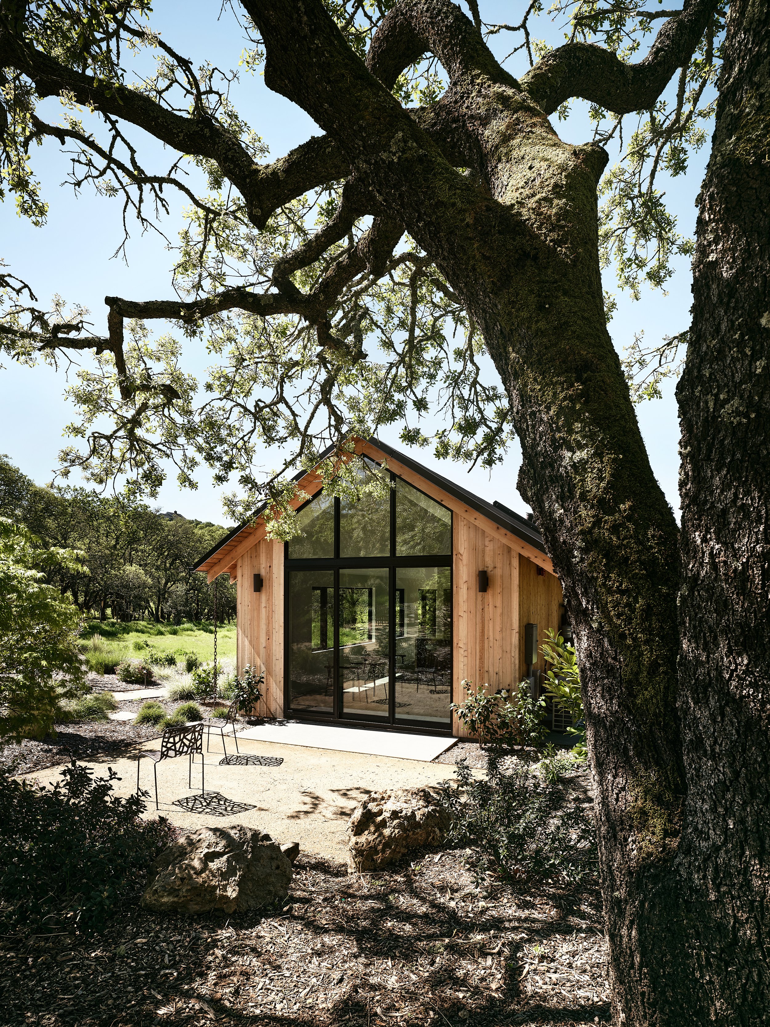 A small modern house with a wooden exterior and large glass front, surrounded by trees and rocks, with outdoor chairs on a sandy ground in the foreground.