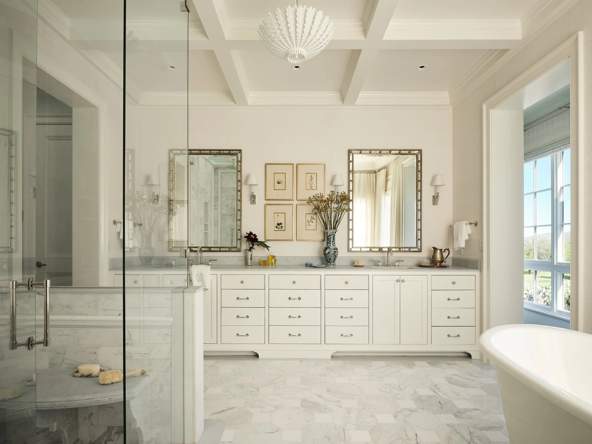 A spacious luxurious bathroom featuring a double vanity with two mirrors, framed art on the wall, a large window with curtains, a freestanding bathtub, and marble flooring.