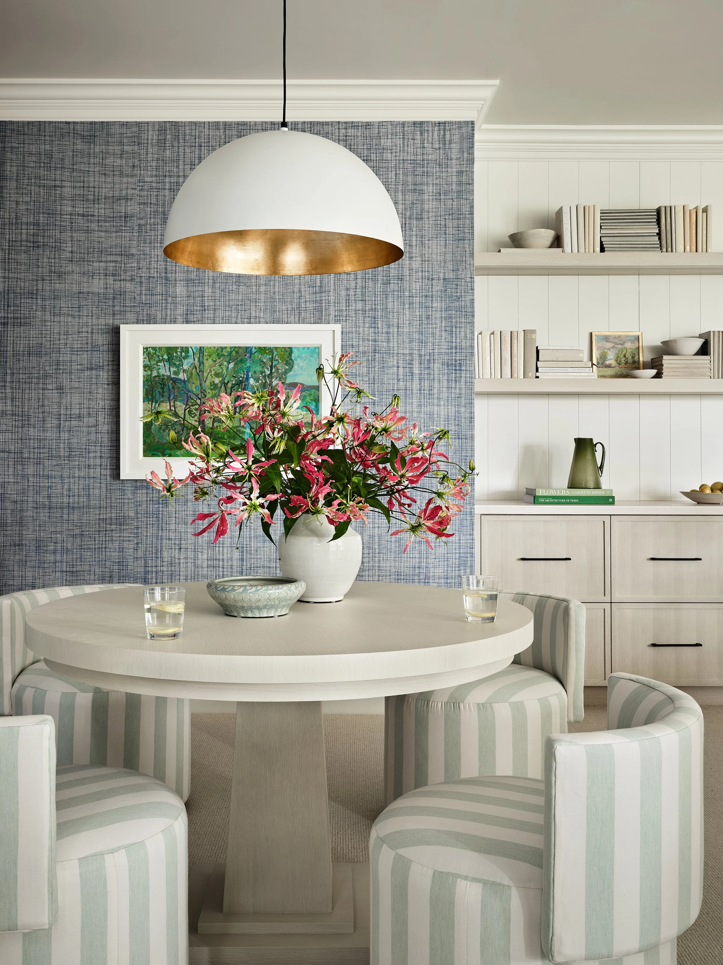 A dining room with a white round table, four striped upholstered chairs, a large white pendant lamp, a colorful flower arrangement in a white vase, a blue textured wall, and built-in white shelves with books, bowls, and decorative items.
