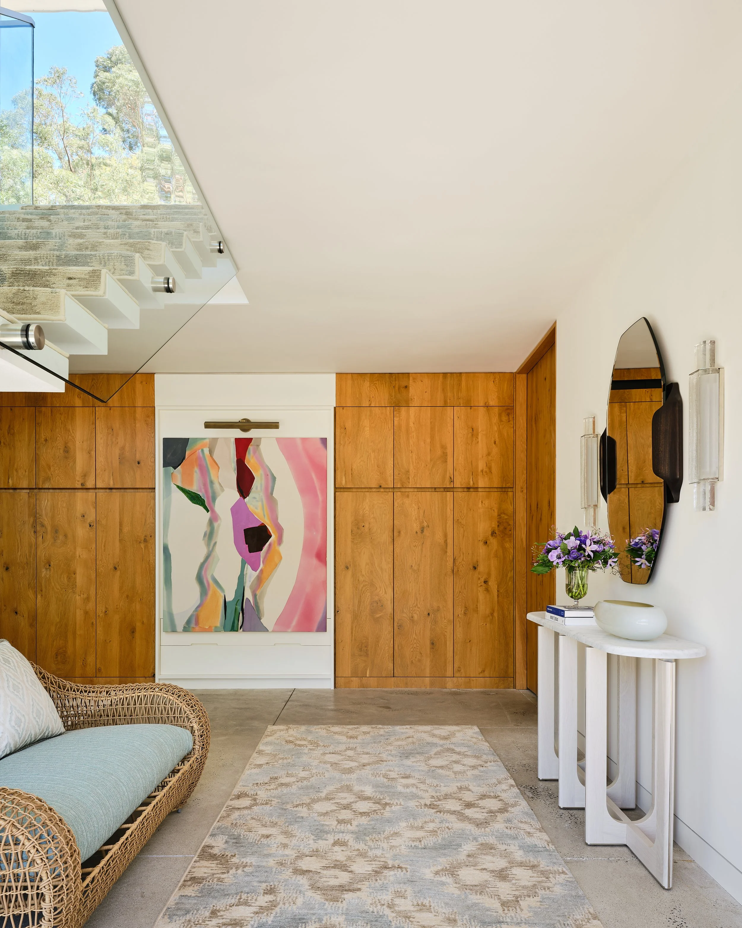 Modern living room with wooden wall panels, abstract artwork, wicker sofa with light blue cushion, and white console table with purple flowers in a glass vase.