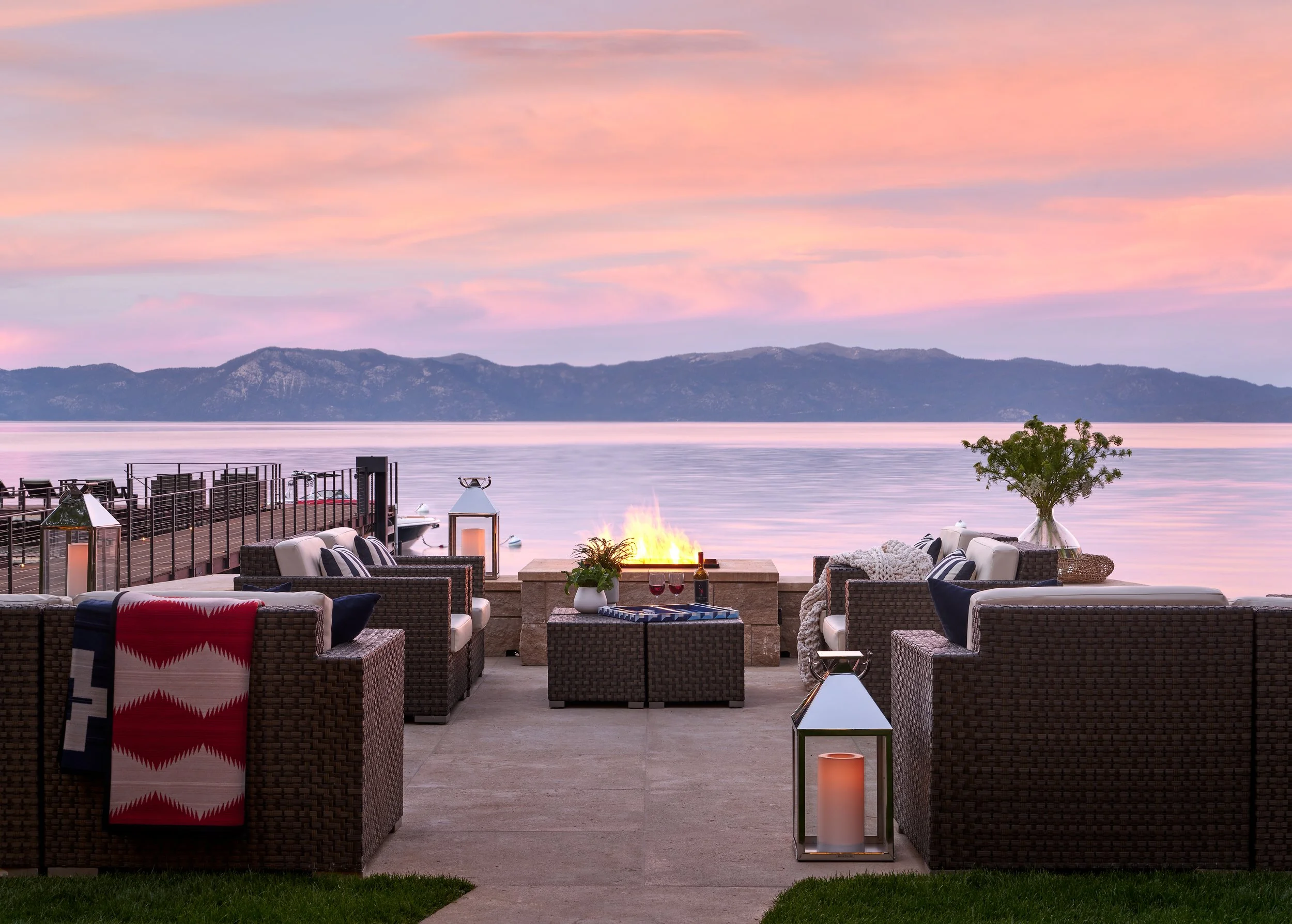 An outdoor patio overlooking a body of water with mountains in the distance during sunset. The patio has wicker chairs, tables with candles, a fire pit, and decorative plants.