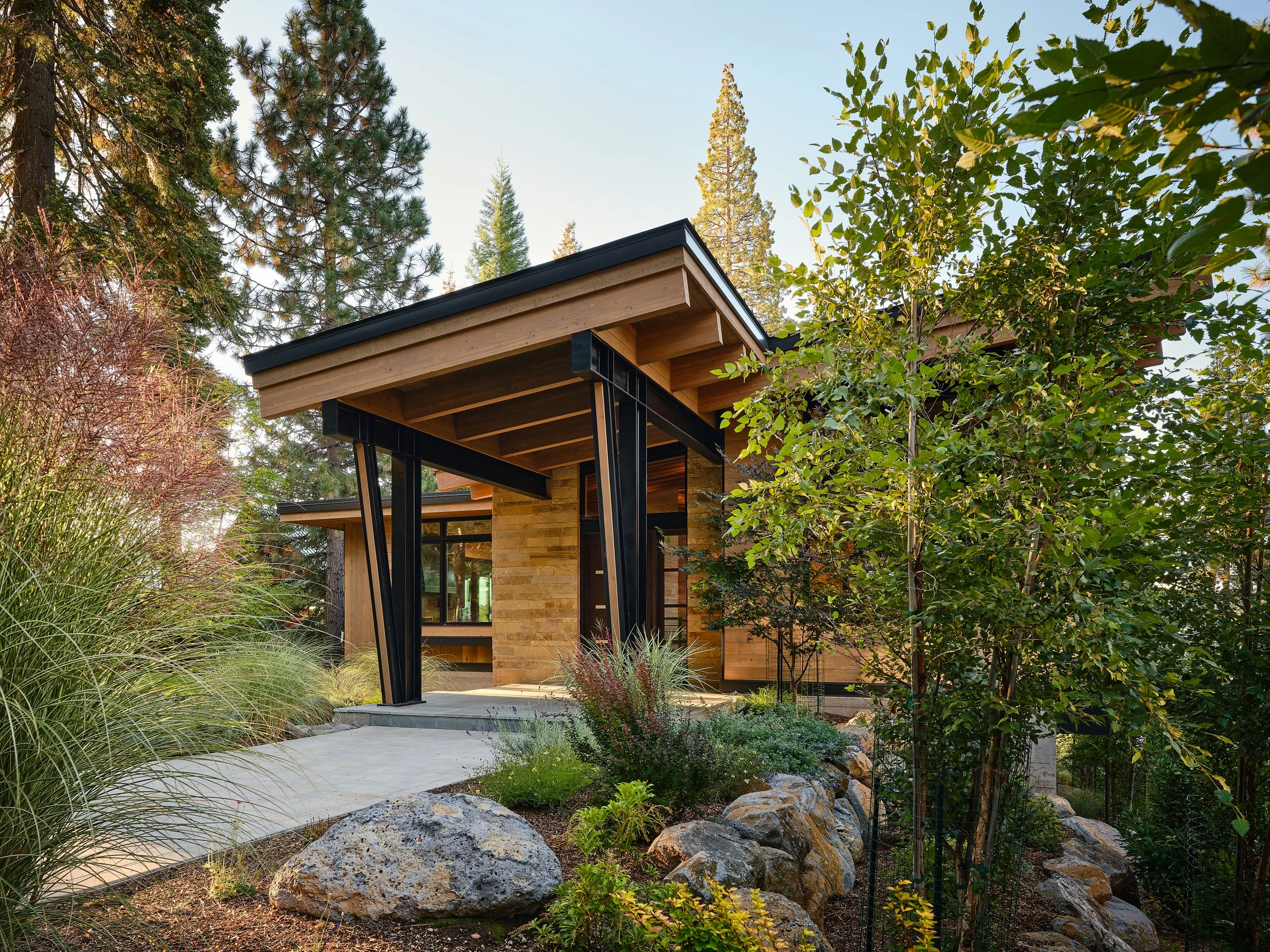Modern house with wooden and stone exterior, surrounded by trees and rocks, with a concrete path leading to the entrance.