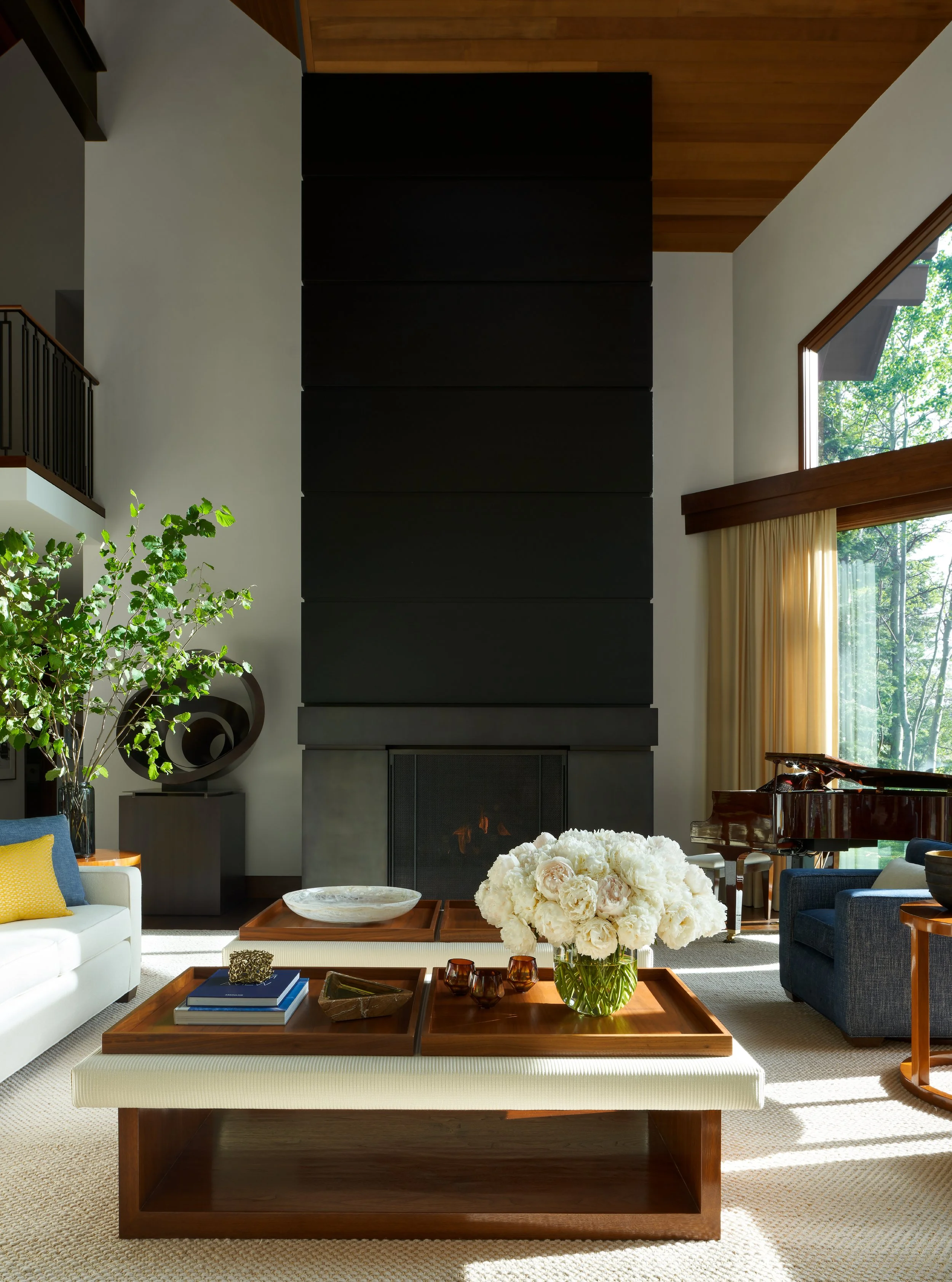 Modern living room with a large black fireplace, white sofa, blue armchair, grand piano, and coffee table with floral centerpiece, natural light.