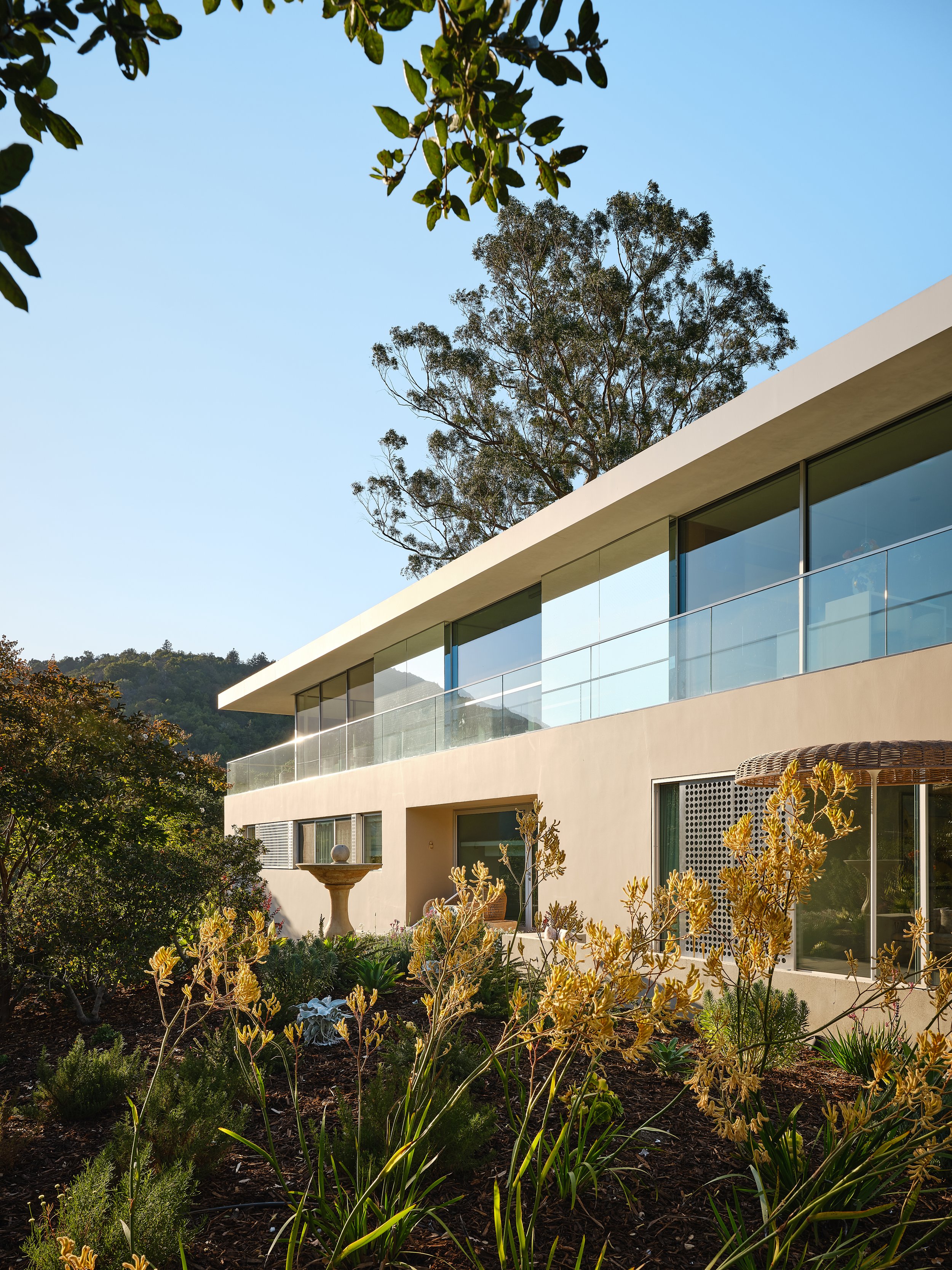 Modern two-story house with large glass windows and a balcony with glass railing, surrounded by garden with plants and a birdbath, trees in the background, clear blue sky overhead.