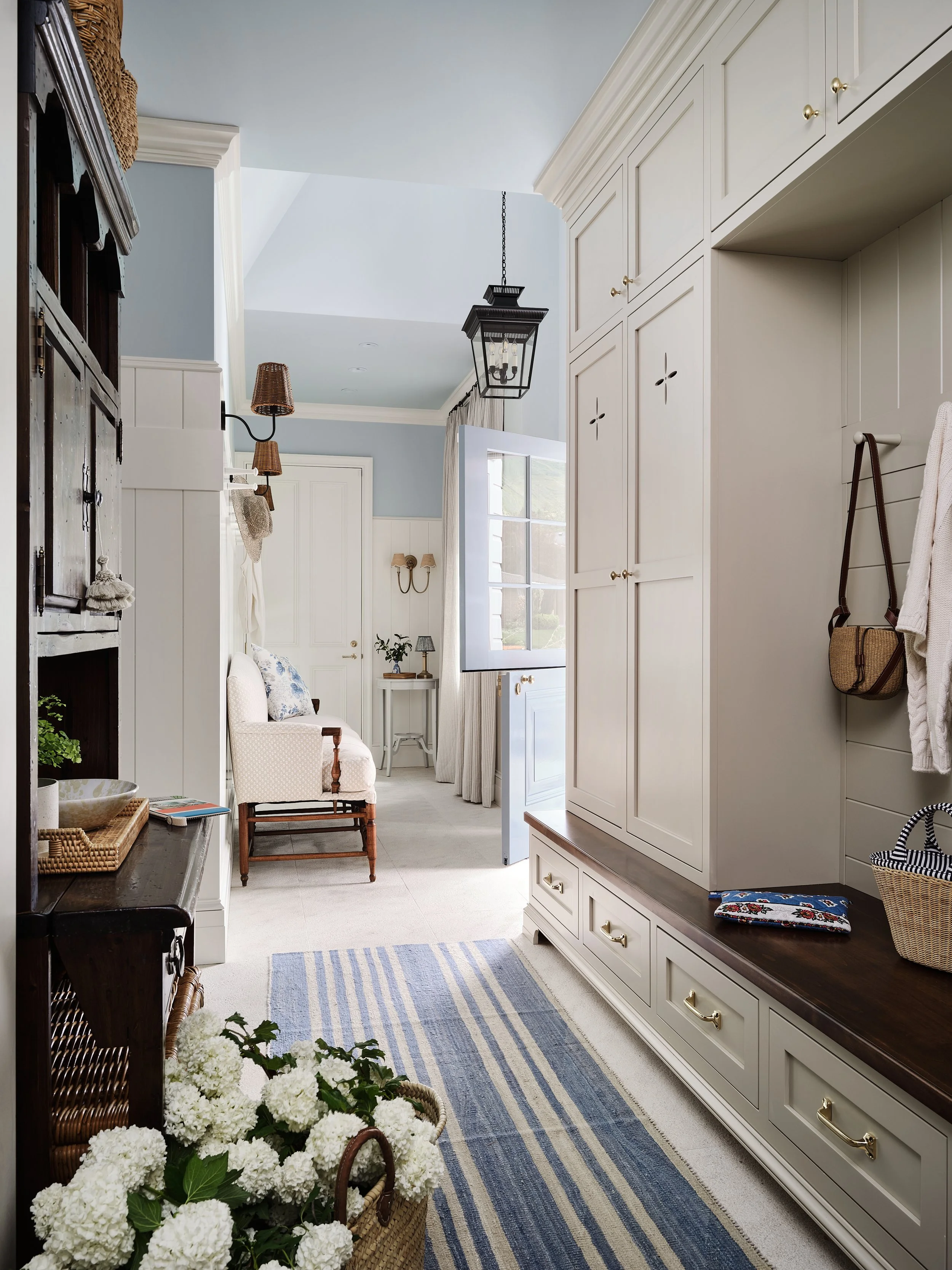 Entryway with white built-in storage, a wooden bench, a large flower basket, and a blue striped rug leading to a sitting area with beige armchair, small side table, and window with curtains.