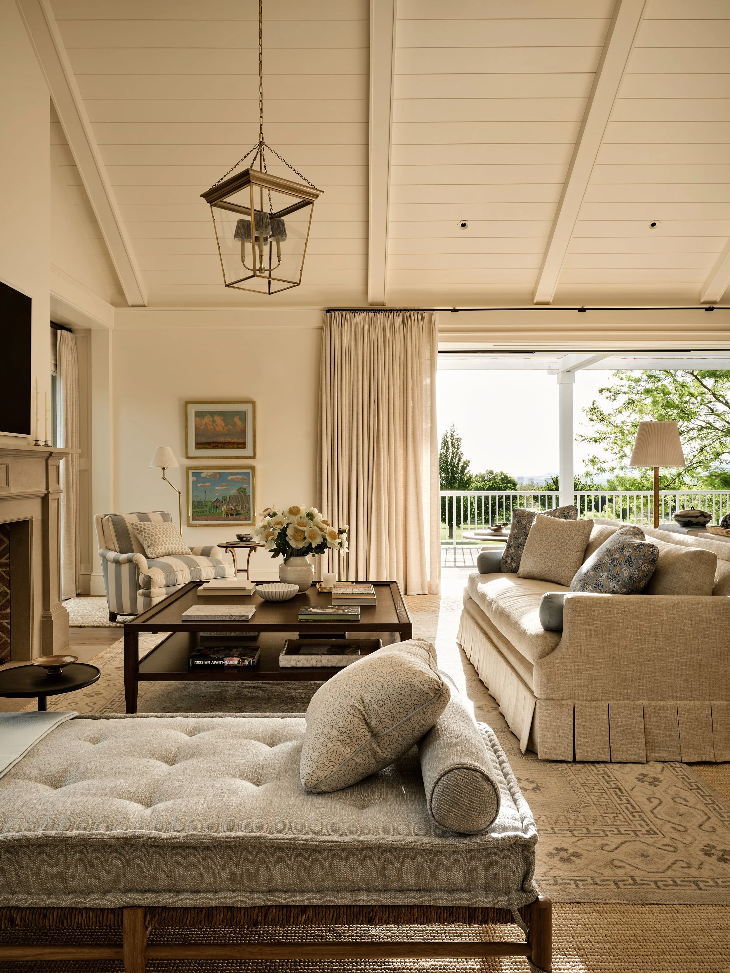 Bright living room with cream-colored furniture, a wood coffee table with books and a flower arrangement, a striped armchair, a large beige sofa with decorative pillows, and a balcony with green trees outside. There are paintings on the wall, a lamp,