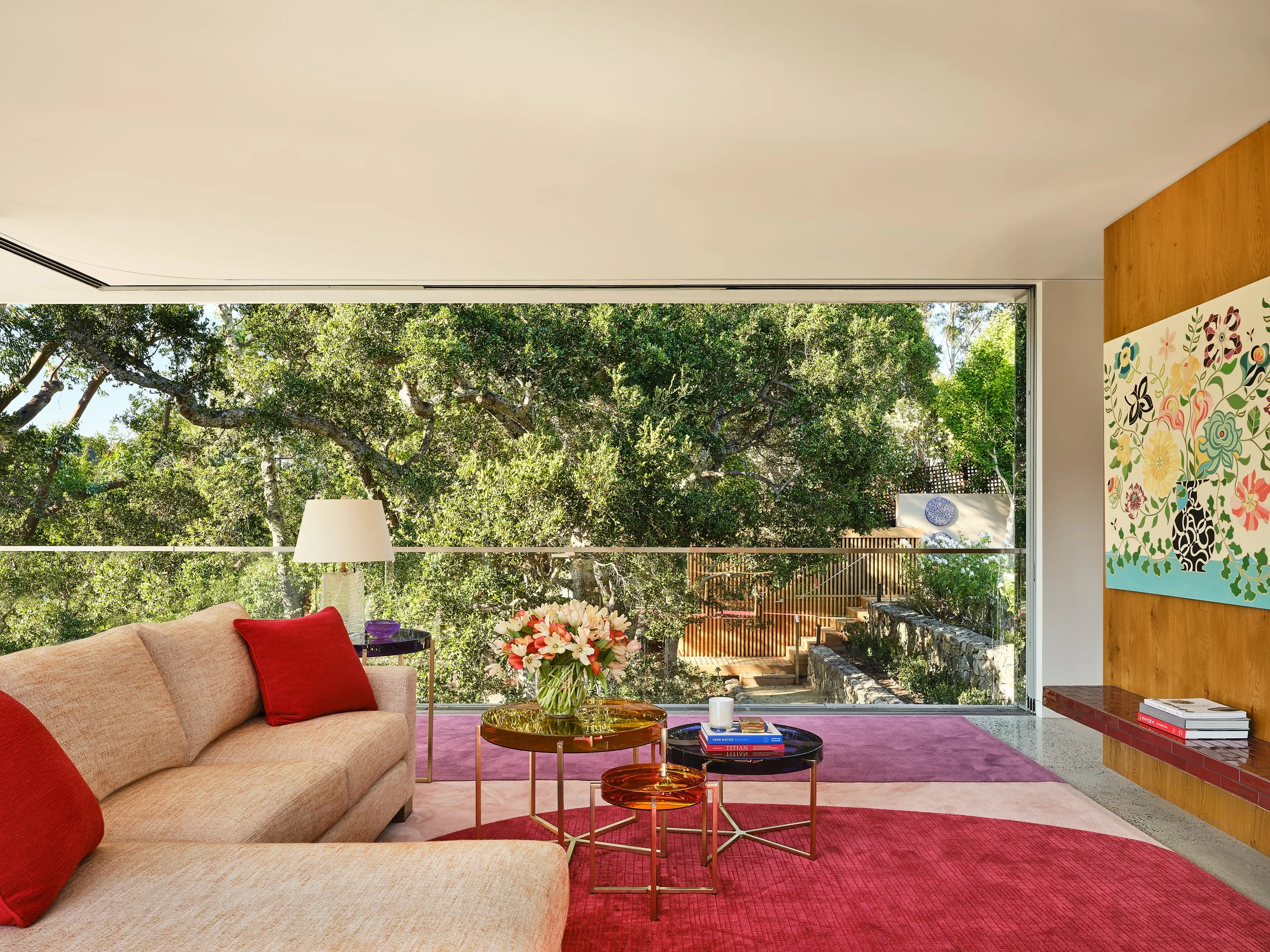 Modern living room with large window overlooking trees, beige sofa with red pillows, colorful nesting tables with flowers, books, and a cup, purple and pink area rugs, painting on wooden wall, and a bench with books.