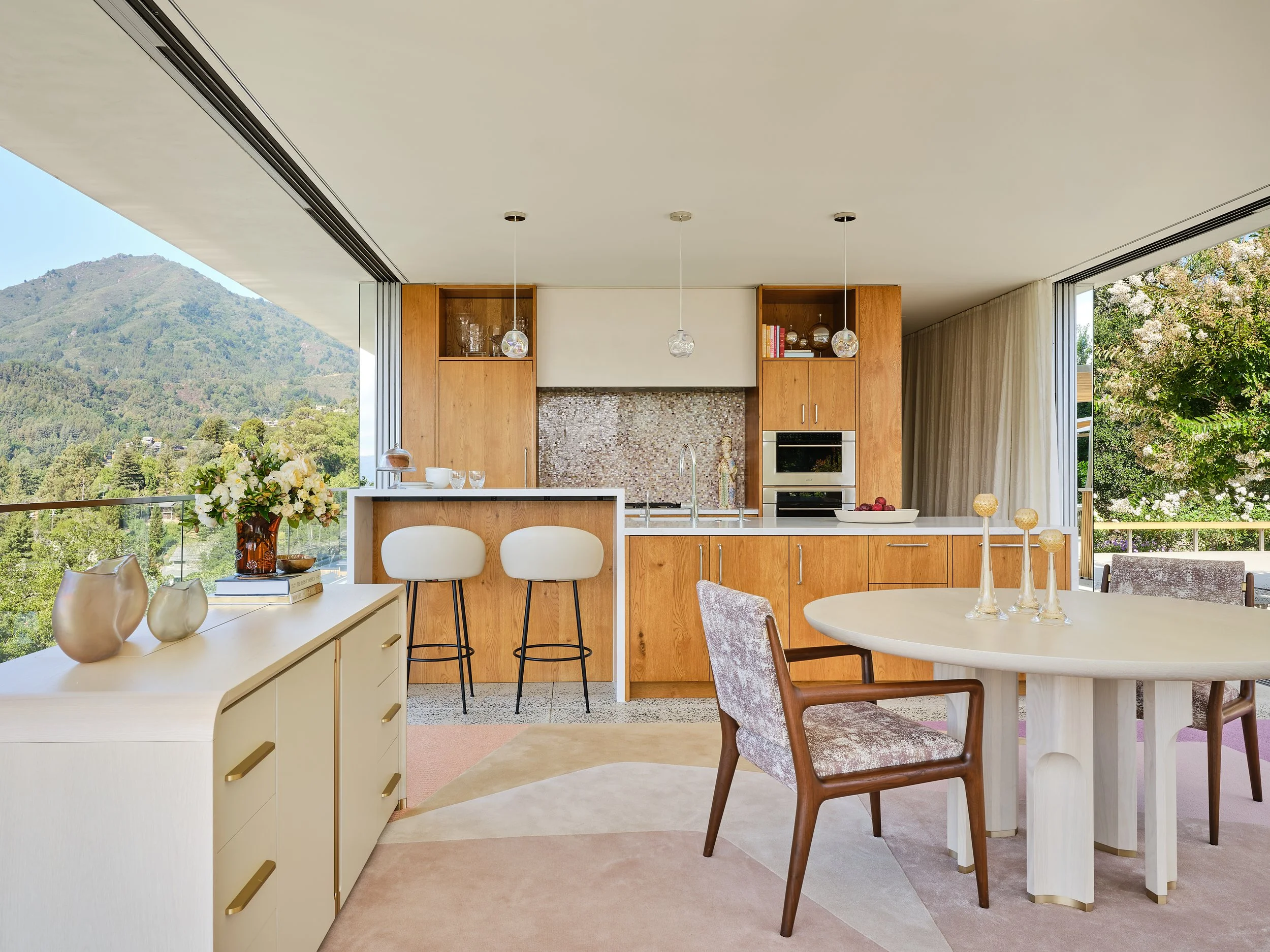 Open-concept kitchen and dining area with large windows showcasing mountain and greenery view, featuring wood cabinetry, a white kitchen island with two stools, a round dining table with chairs, and decorative vases and flowers.