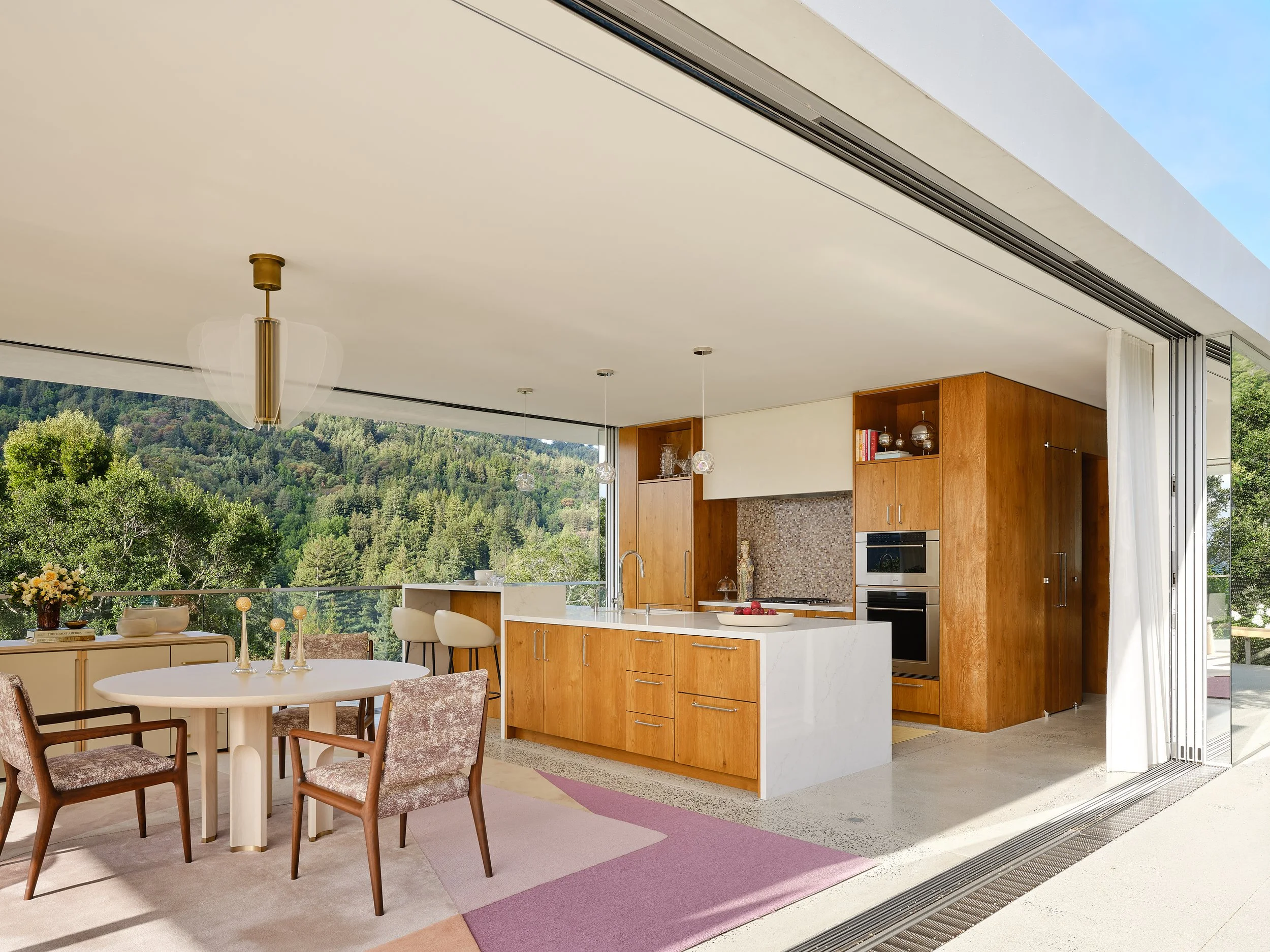 Contemporary open-plan kitchen and dining area with floor-to-ceiling windows showing a green mountainous view. The space includes a white round dining table with four upholstered chairs, a kitchen island with a sink, and built-in wooden cabinets with
