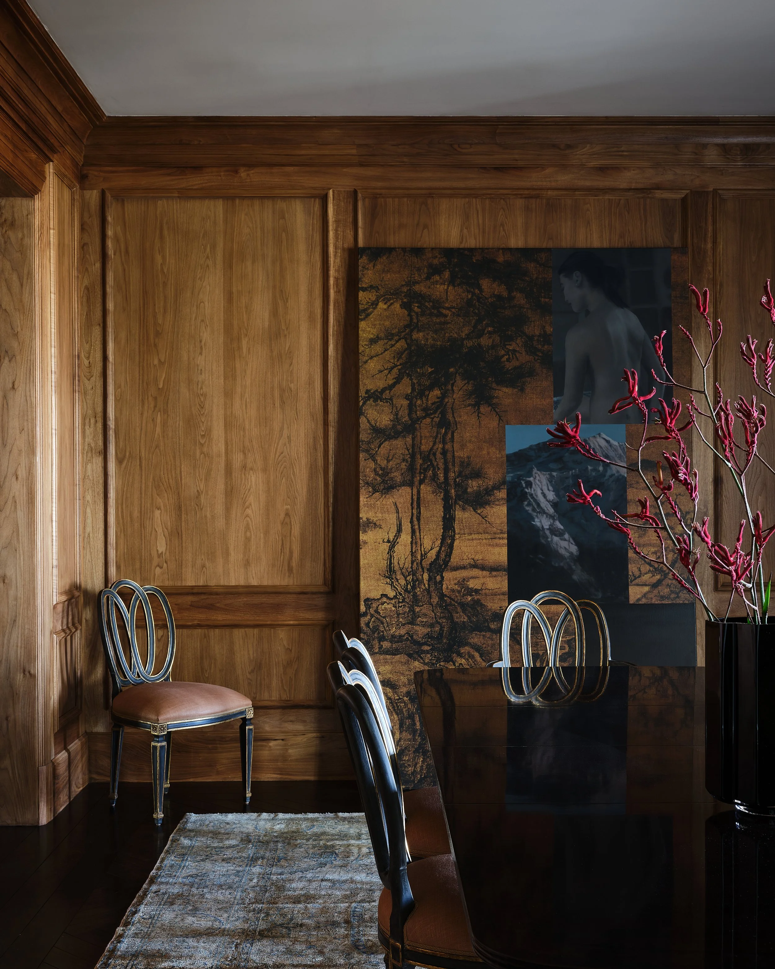 A dining room with wood-paneled walls and a dark wooden table. Four chairs are visible, three with black frames and brown seats, and one with a black and gold frame and pink cushion. A large black vase with red branches and flowers stands on the righ