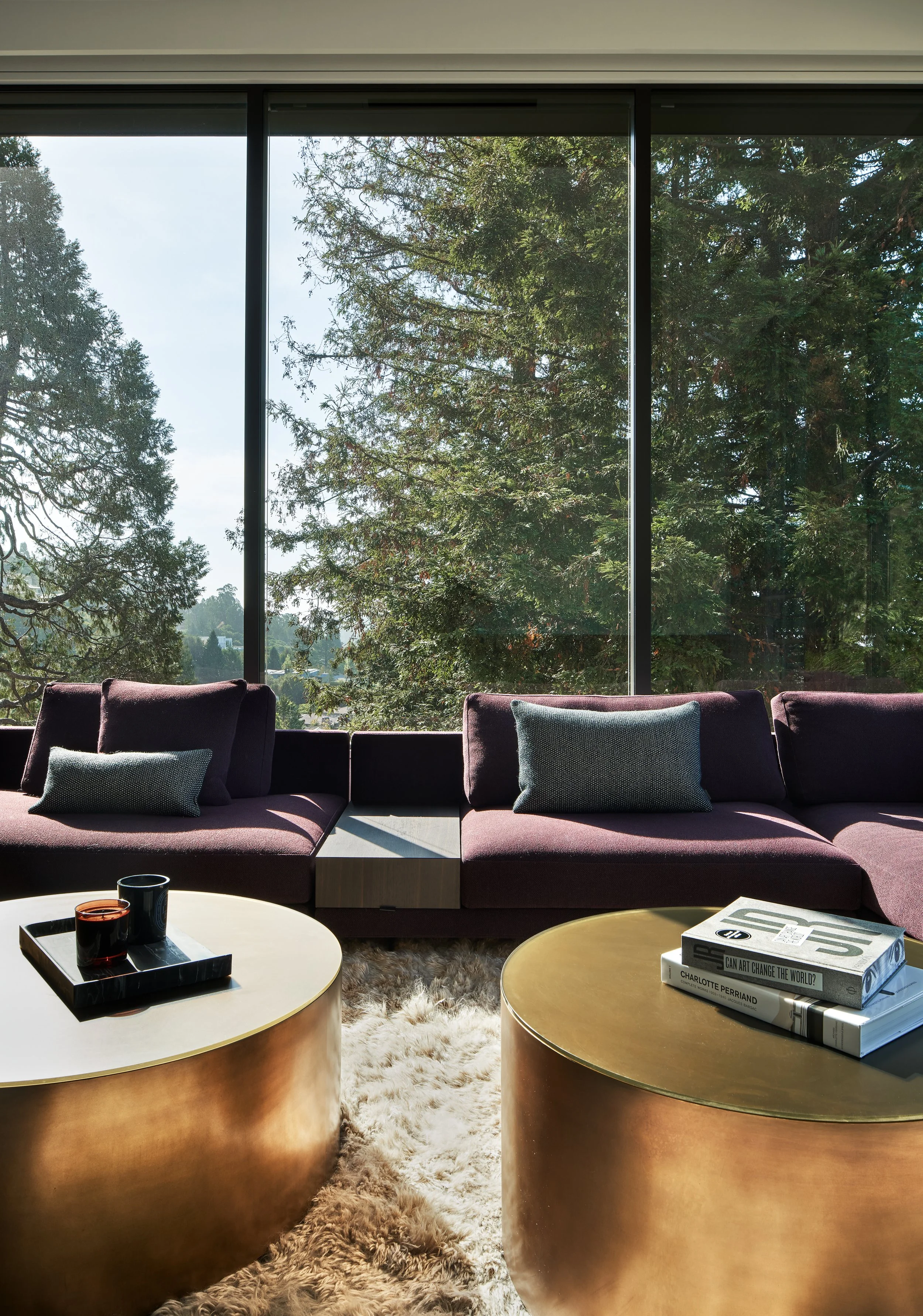 Living room with large windows showing trees outside, purple sofa with cushions, two round metallic coffee tables, a fuzzy rug, and books on one of the tables.