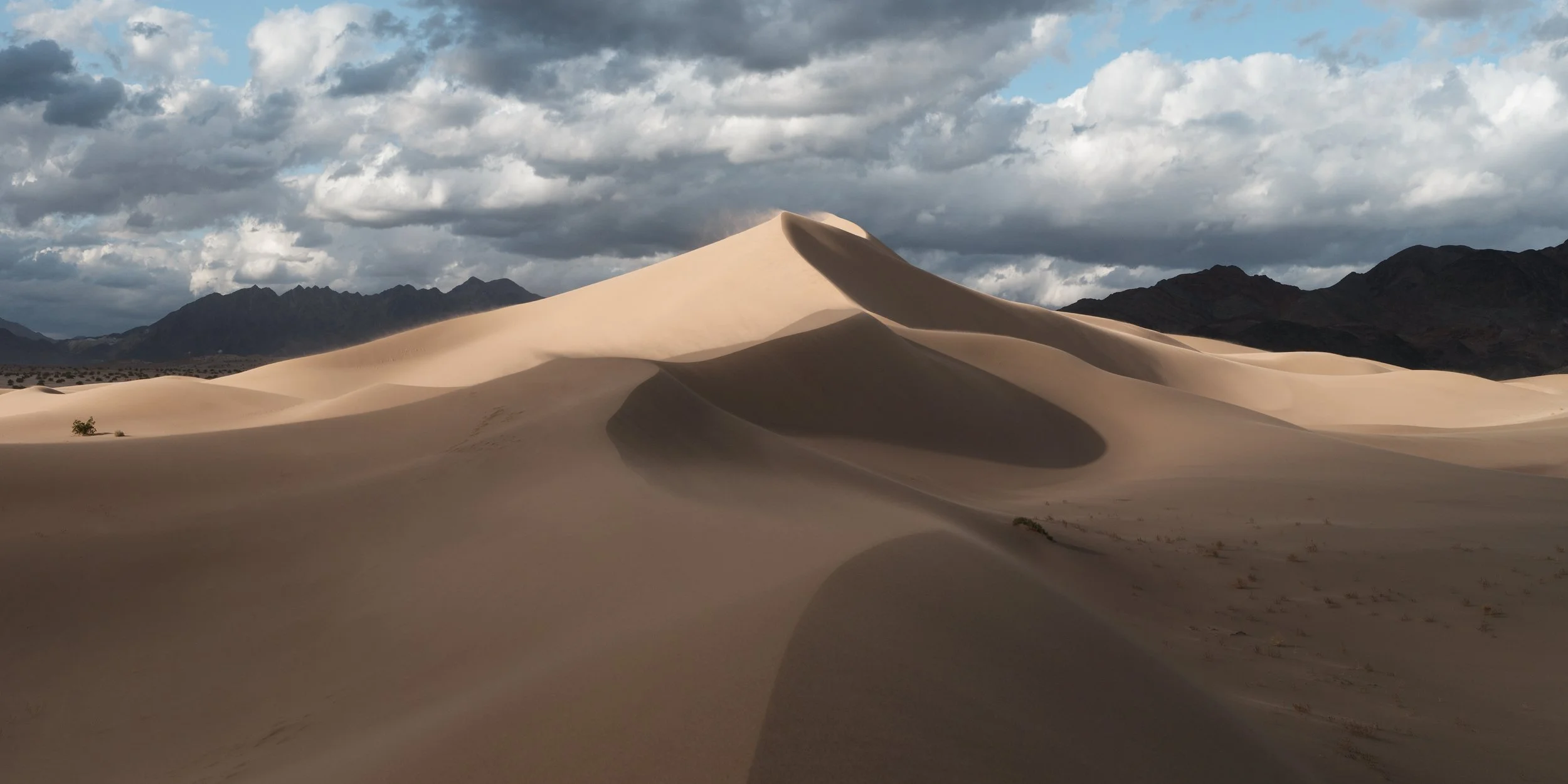Sand dunes in a desert with mountains and cloudy sky in the background.