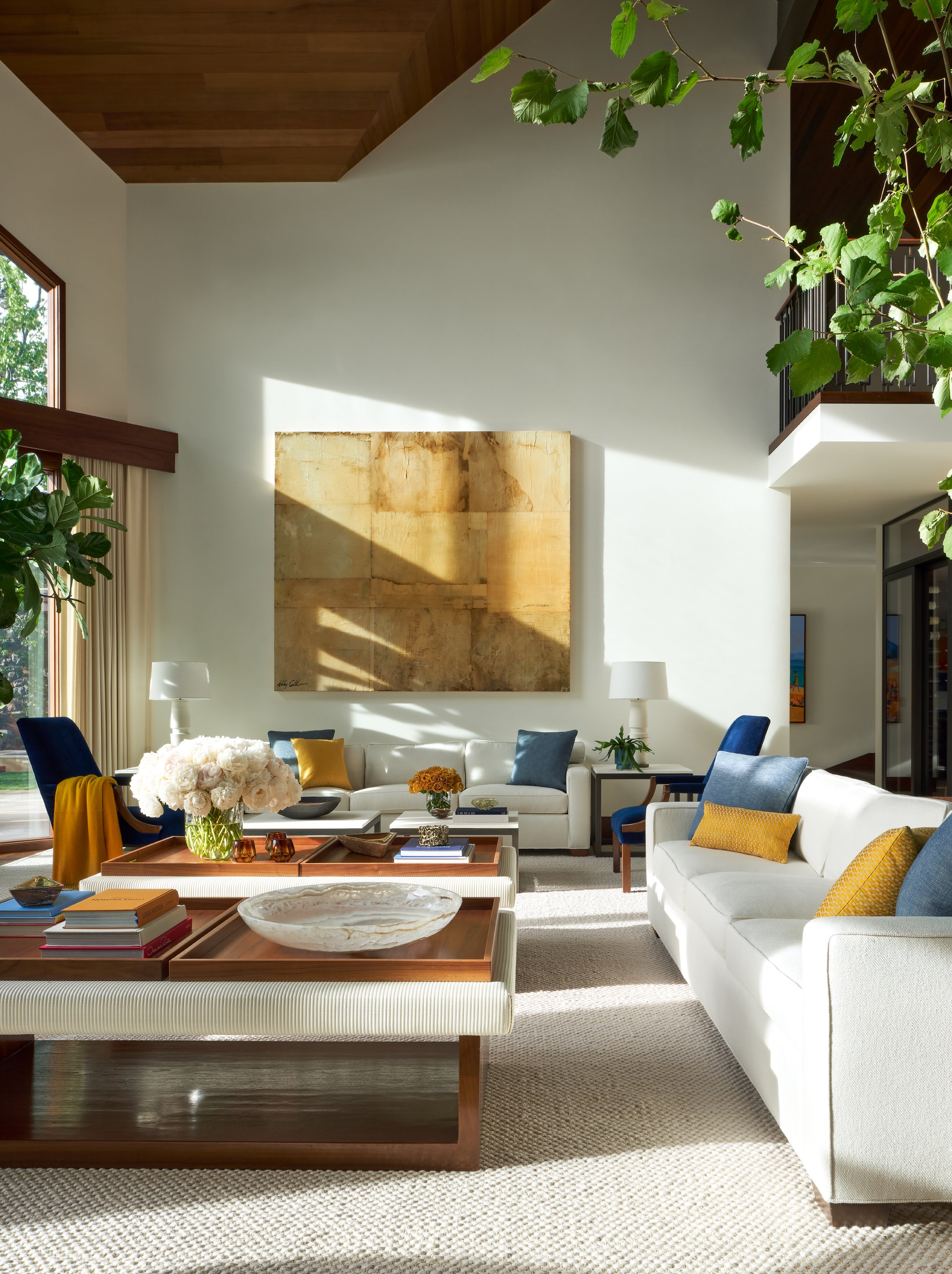 Bright living room with white sofa, blue and yellow accent pillows, large abstract artwork, and a coffee table with books and decorative items, illuminated by natural light through large windows.