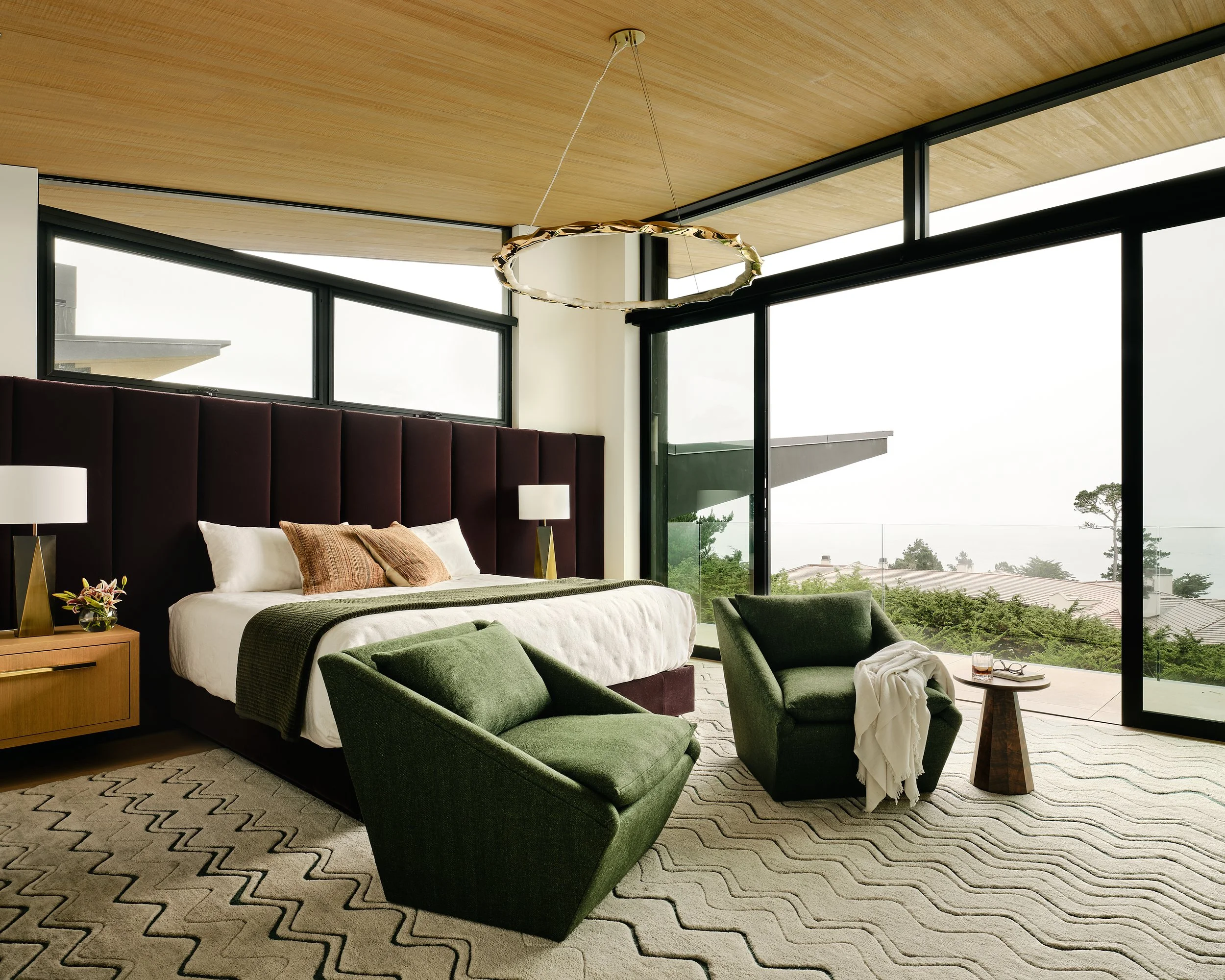 Modern bedroom with large windows, green armchairs, a bed with a dark headboard, white bedding, and a patterned rug.
