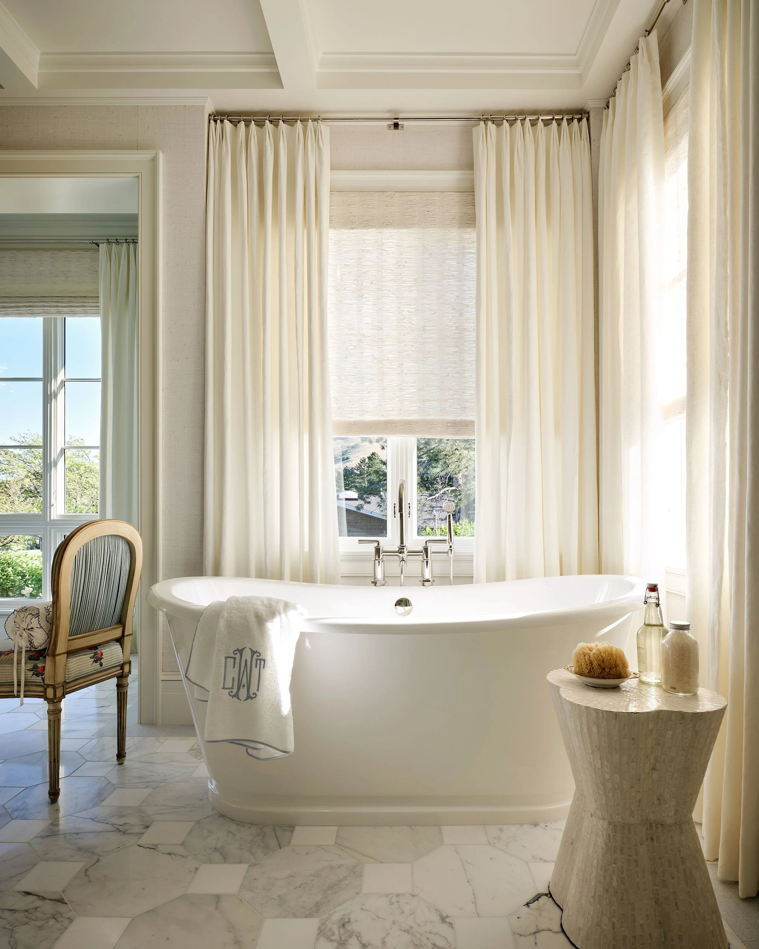 Bright bathroom featuring a white bathtub with a monogrammed towel, surrounded by cream-colored curtains and marble-tile flooring, with a small side table holding a sponge and bottles.