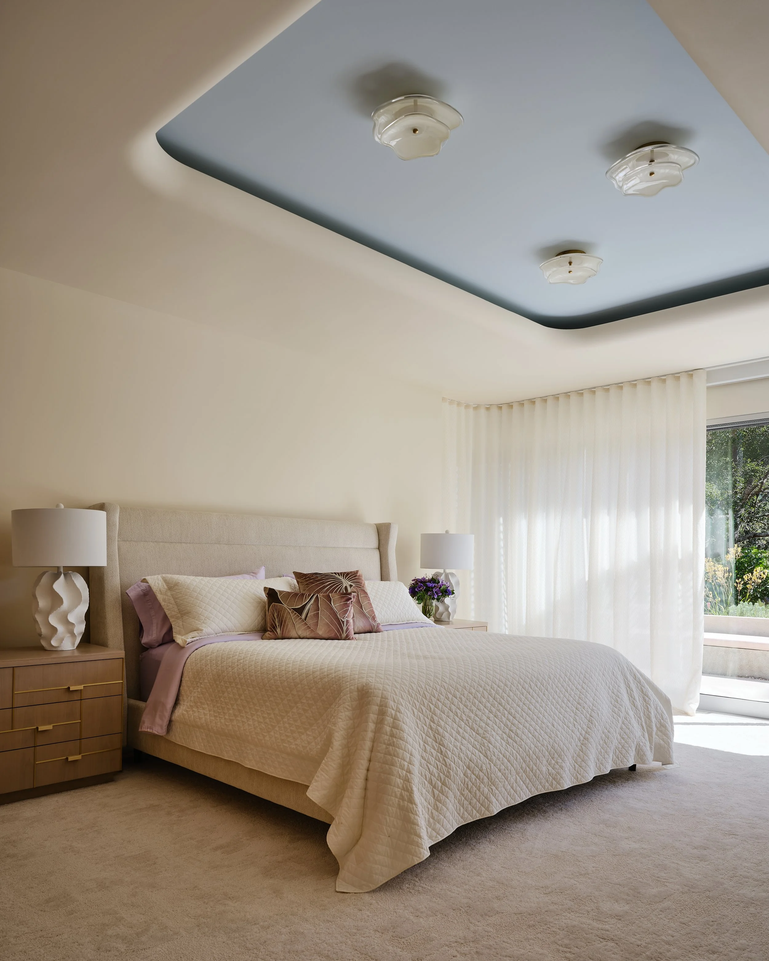 A bedroom with a beige bed, matching pillows, nightstands with lamps, a window with white curtains, and a ceiling with blue paint and white ceiling fixtures.