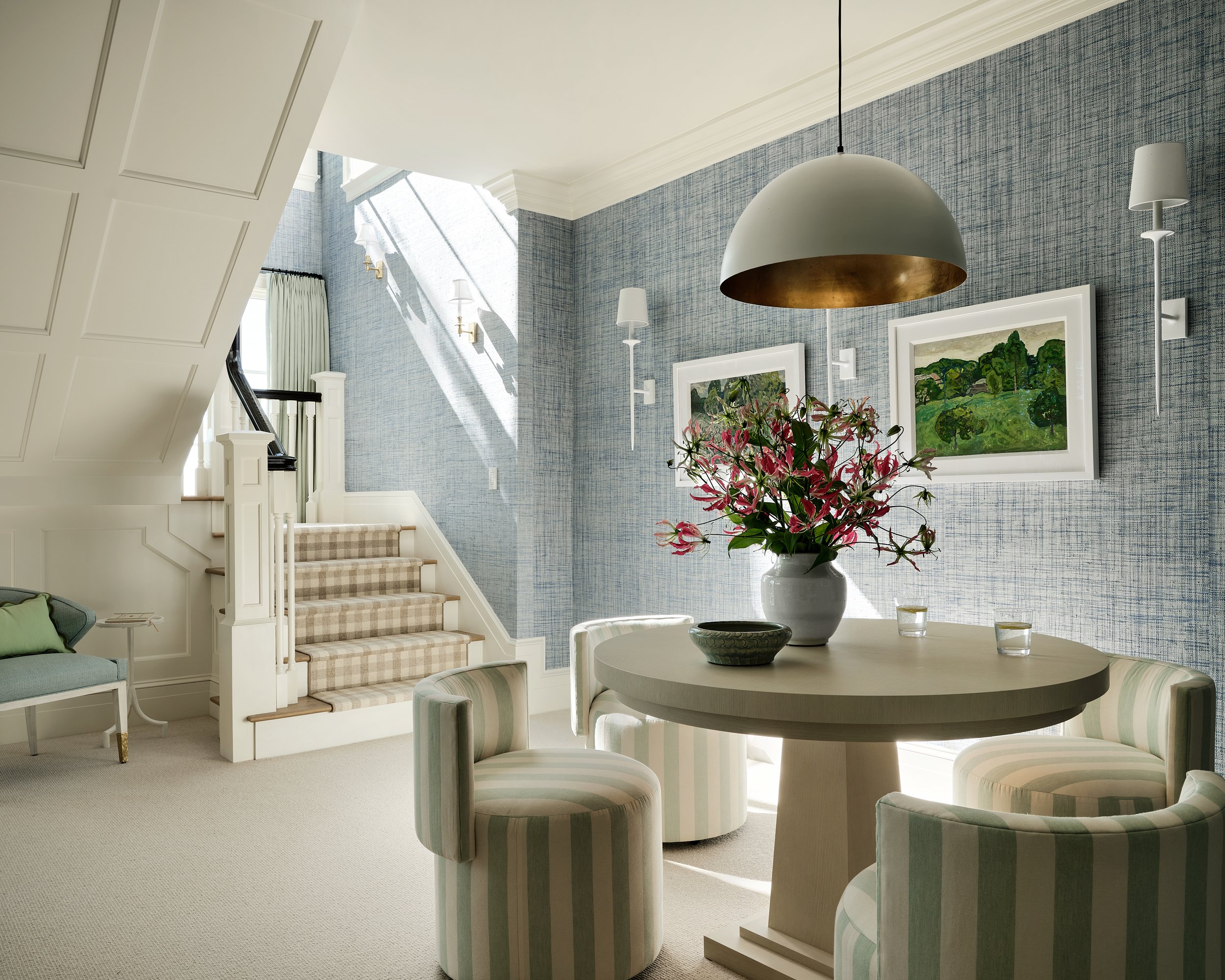 Bright living room with staircase, round table with striped chairs, flower arrangement, wall art, and blue patterned wallpaper.