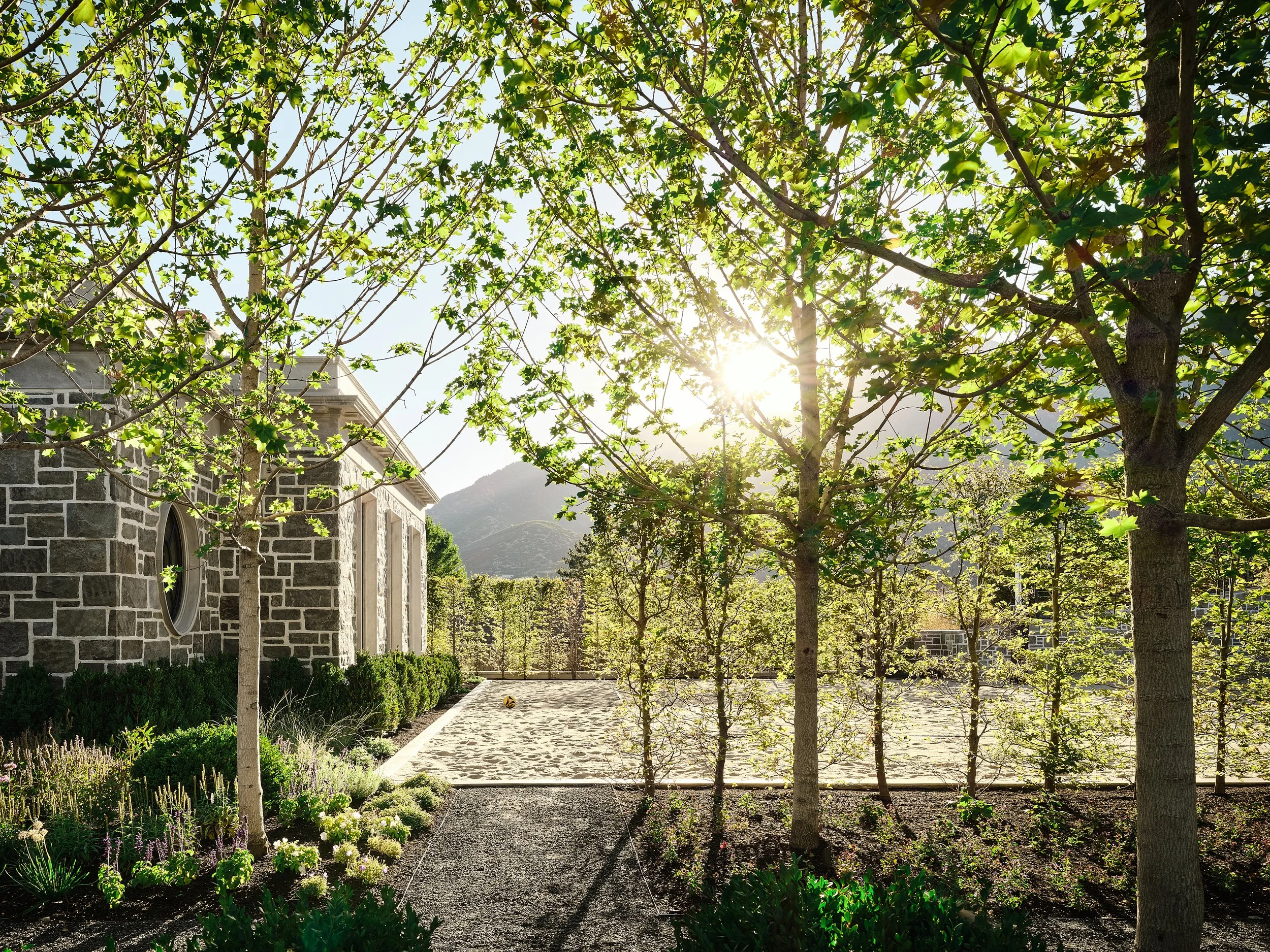 Sunlit garden with trees, bushes, and a stone building on the left, with mountains in the background and a sandy area.