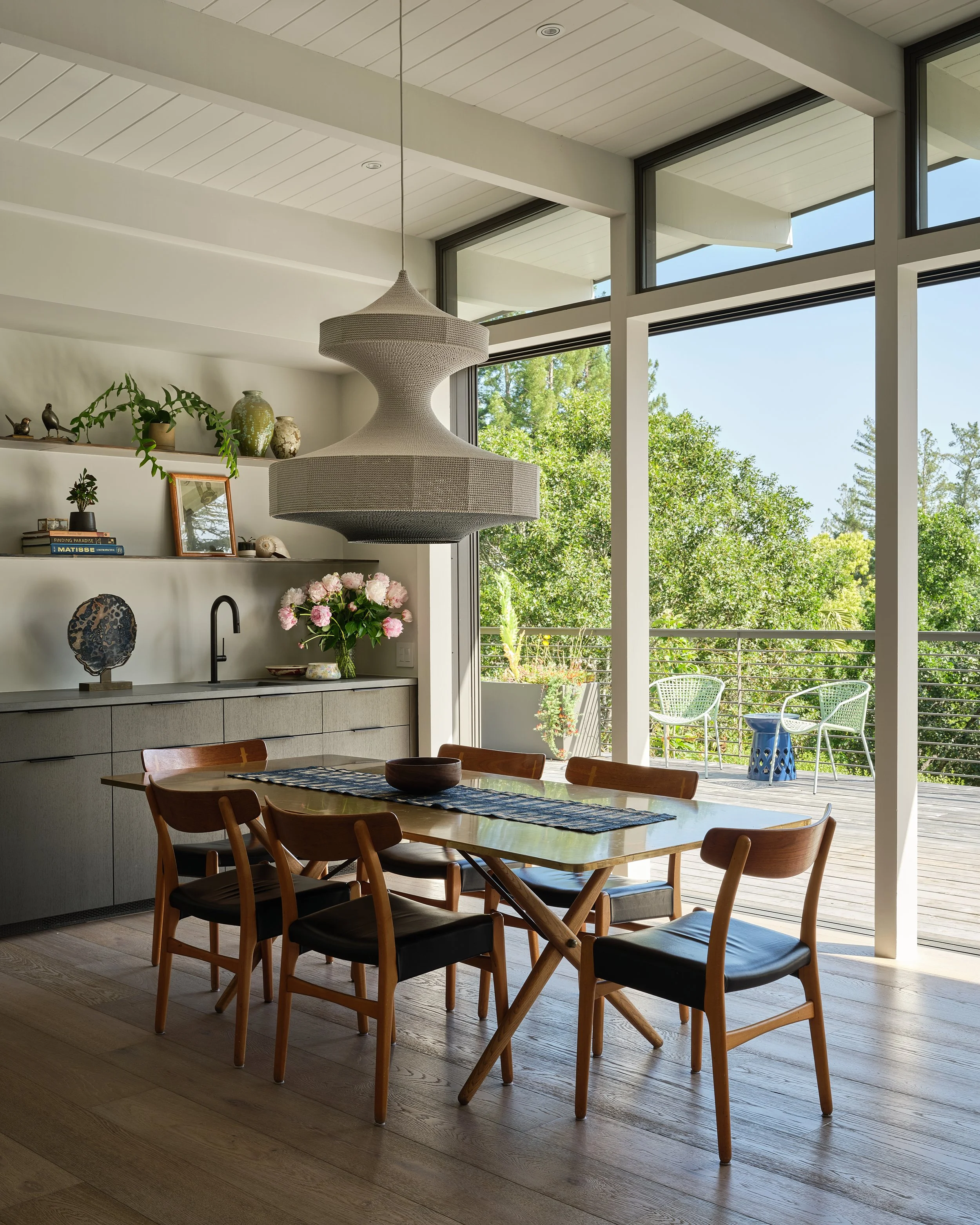 A modern dining room with a wooden table and six chairs, a large pendant light, a sideboard with decorative items, a vase of pink flowers, and a view of a balcony with two chairs and trees outside.