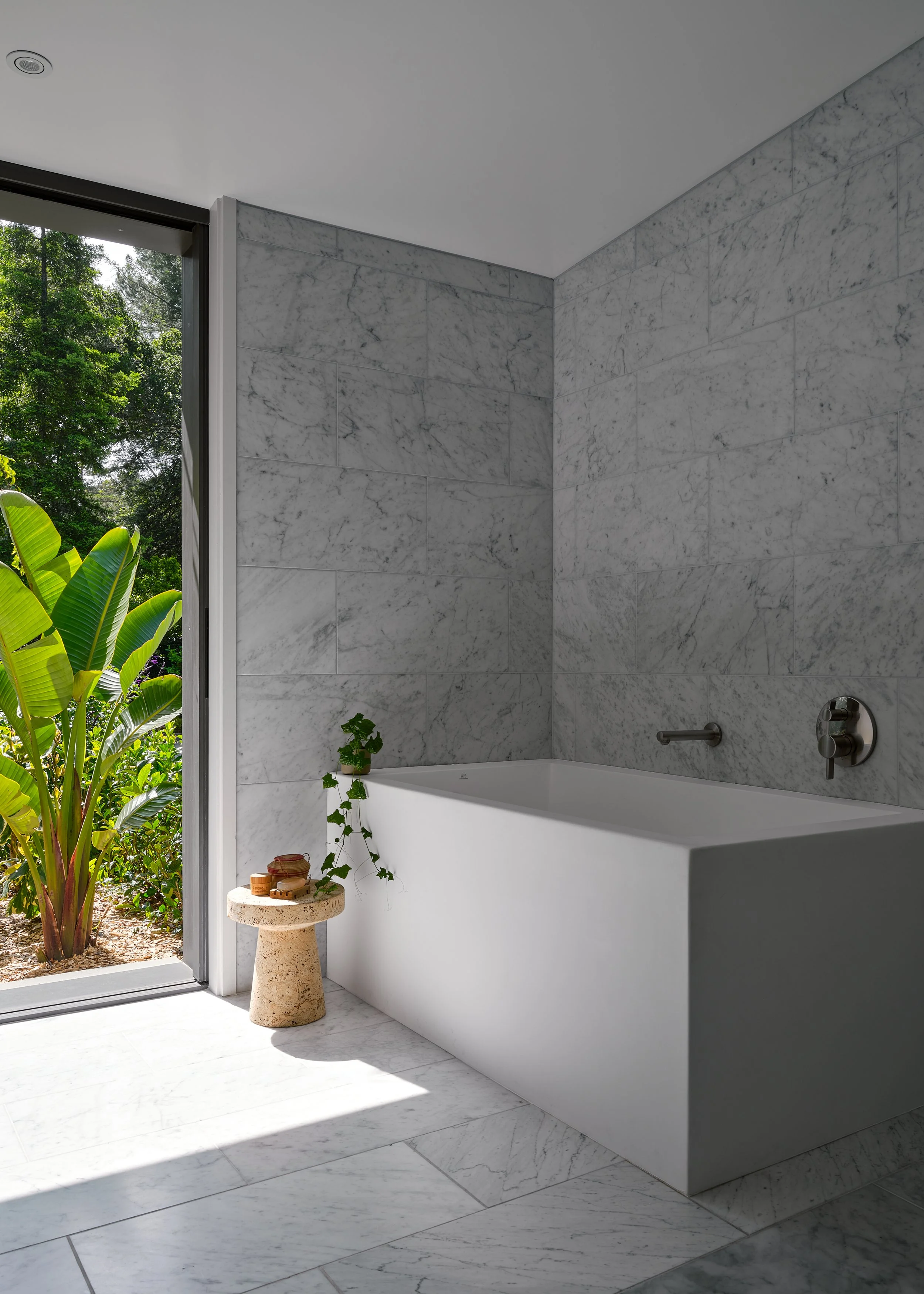 Modern bathroom with a white bathtub, gray marble walls and flooring, and a large open window showing green foliage outside.