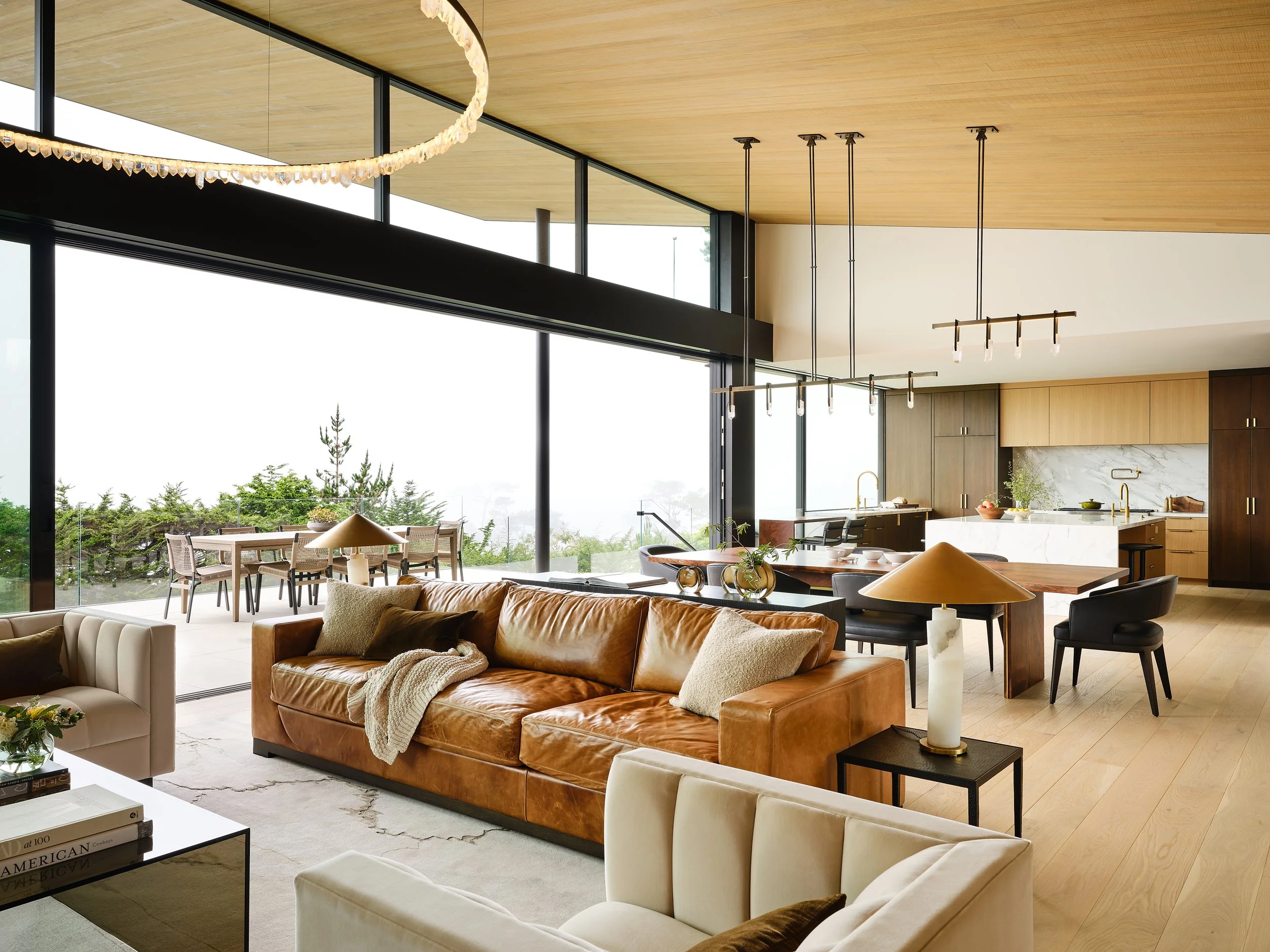 Modern open-concept living room and kitchen with large floor-to-ceiling windows, tan leather sofa, white armchair, wooden dining table, and minimalist chandelier lighting.
