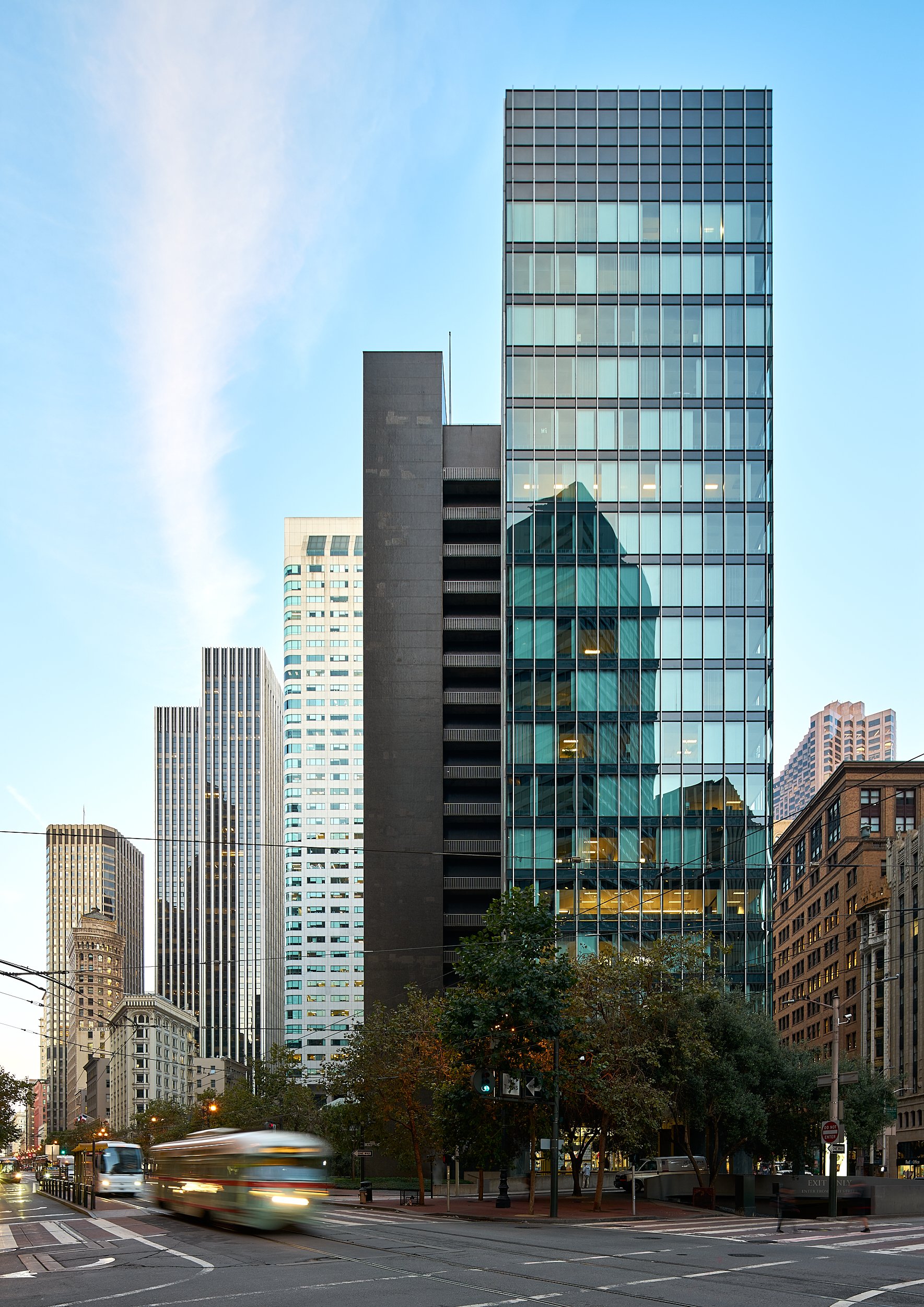 City street scene with tall modern glass and concrete skyscrapers, trees, and moving vehicles at dusk.