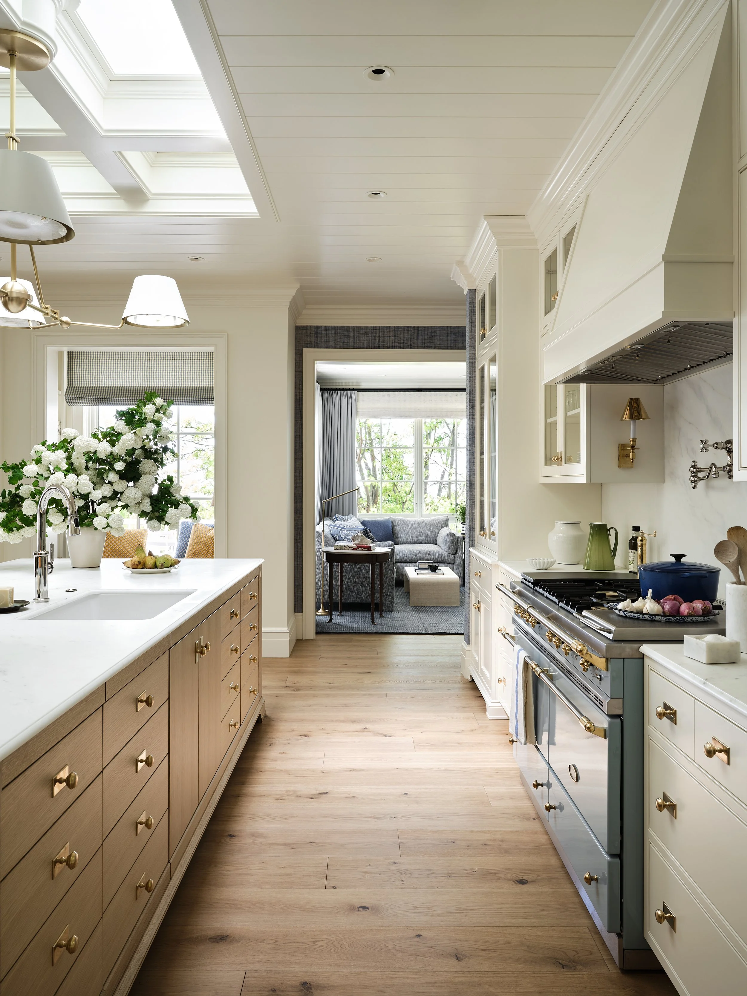 Bright kitchen with white cabinetry, light wood flooring, a gas stove with brass handles, a white countertop with a sink, and a view into a sitting area with large windows and greenery outside.