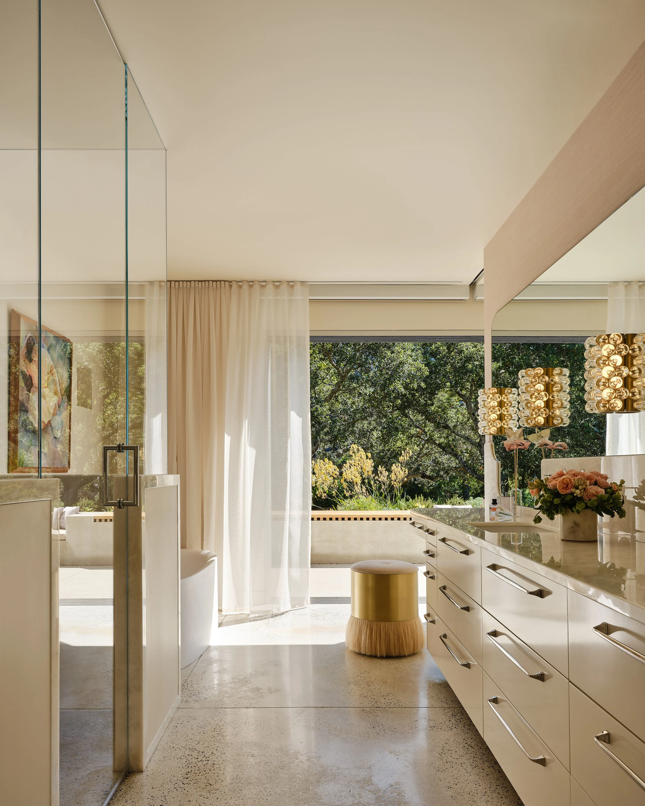 Bright bathroom with white cabinetry, a gold and fur-stool, floral arrangement, and large window with greenery outside.