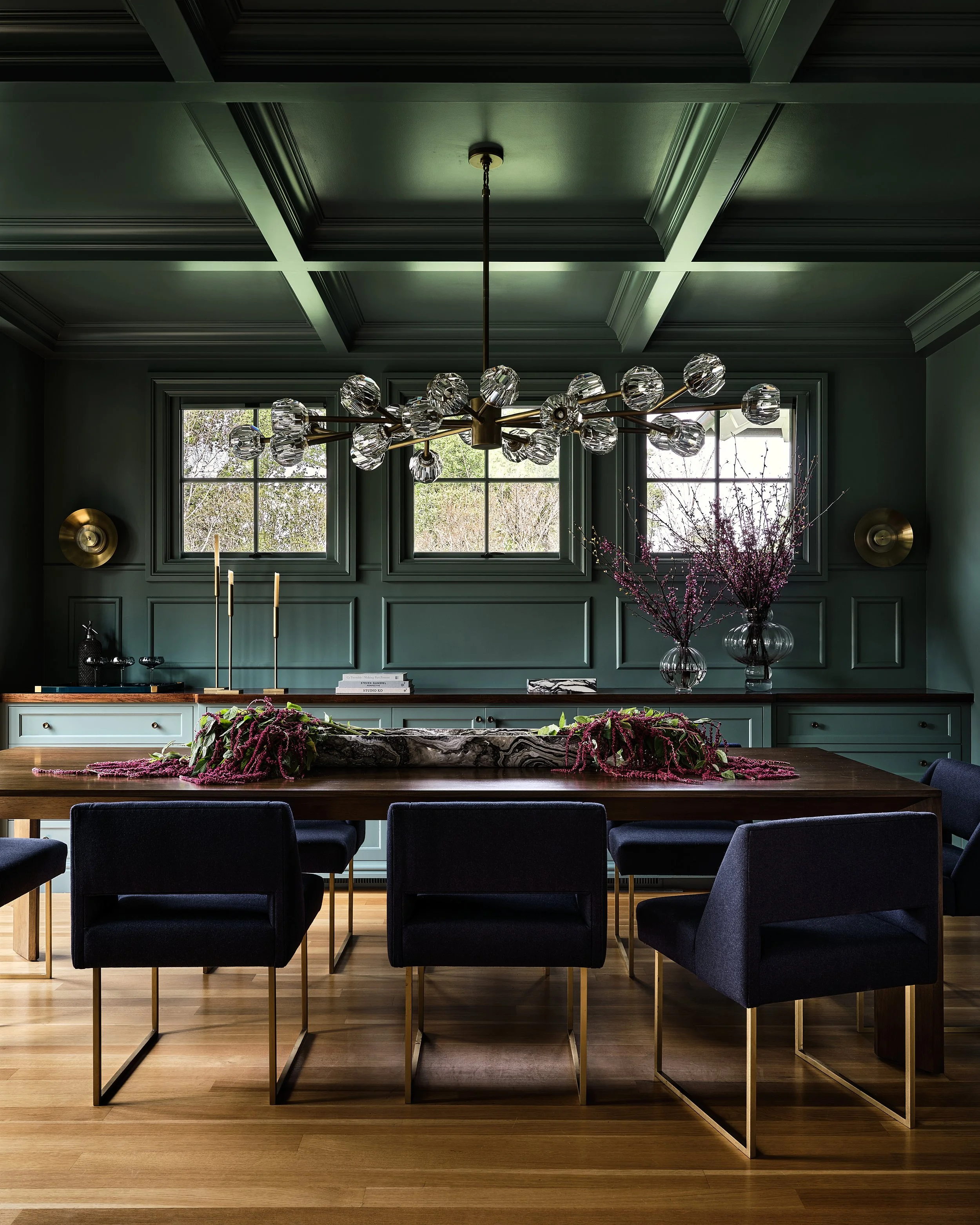 A dark green dining room with a wooden table, black upholstered chairs with gold legs, a modern chandelier with glass globes, vases with pink flowering branches, and windows showing trees outside.