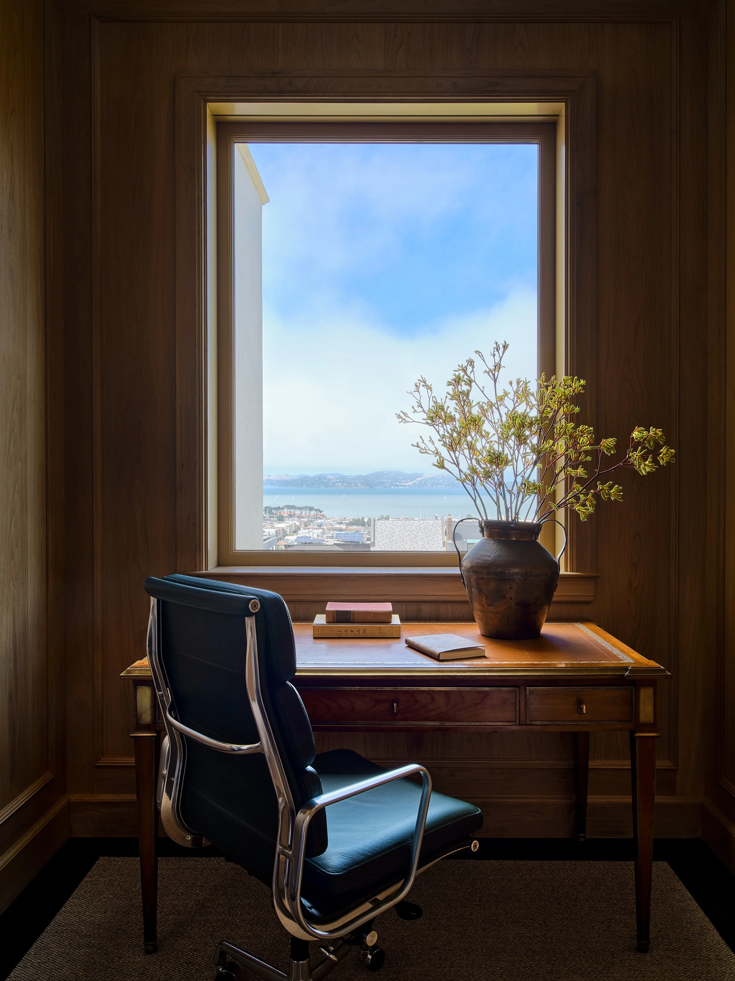 A wooden desk with a potted plant and books sits near a large window overlooking a cityscape and body of water, with a black office chair in front.