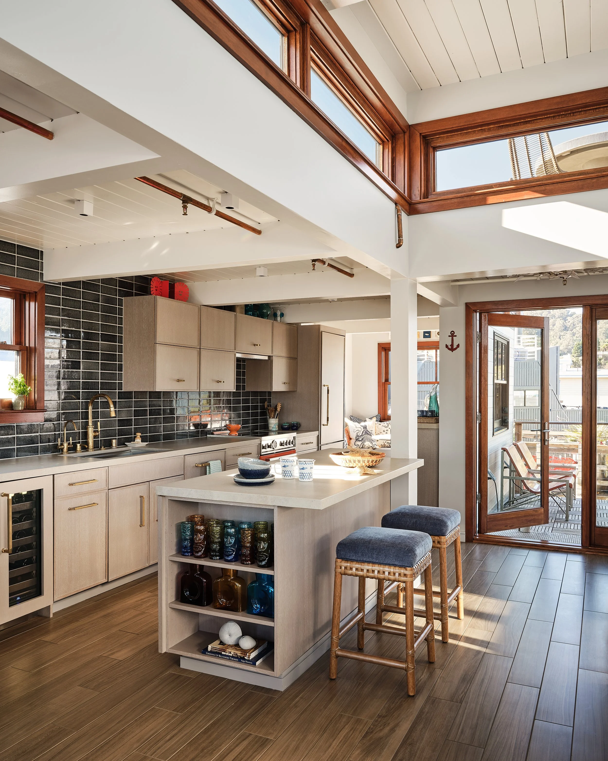 Modern kitchen with light wood cabinets, black tile backsplash, and a kitchen island with colorful glassware and bowls. There are two stools with blue cushions at the island, and large windows and a glass door with wood trim letting in sunlight. An o