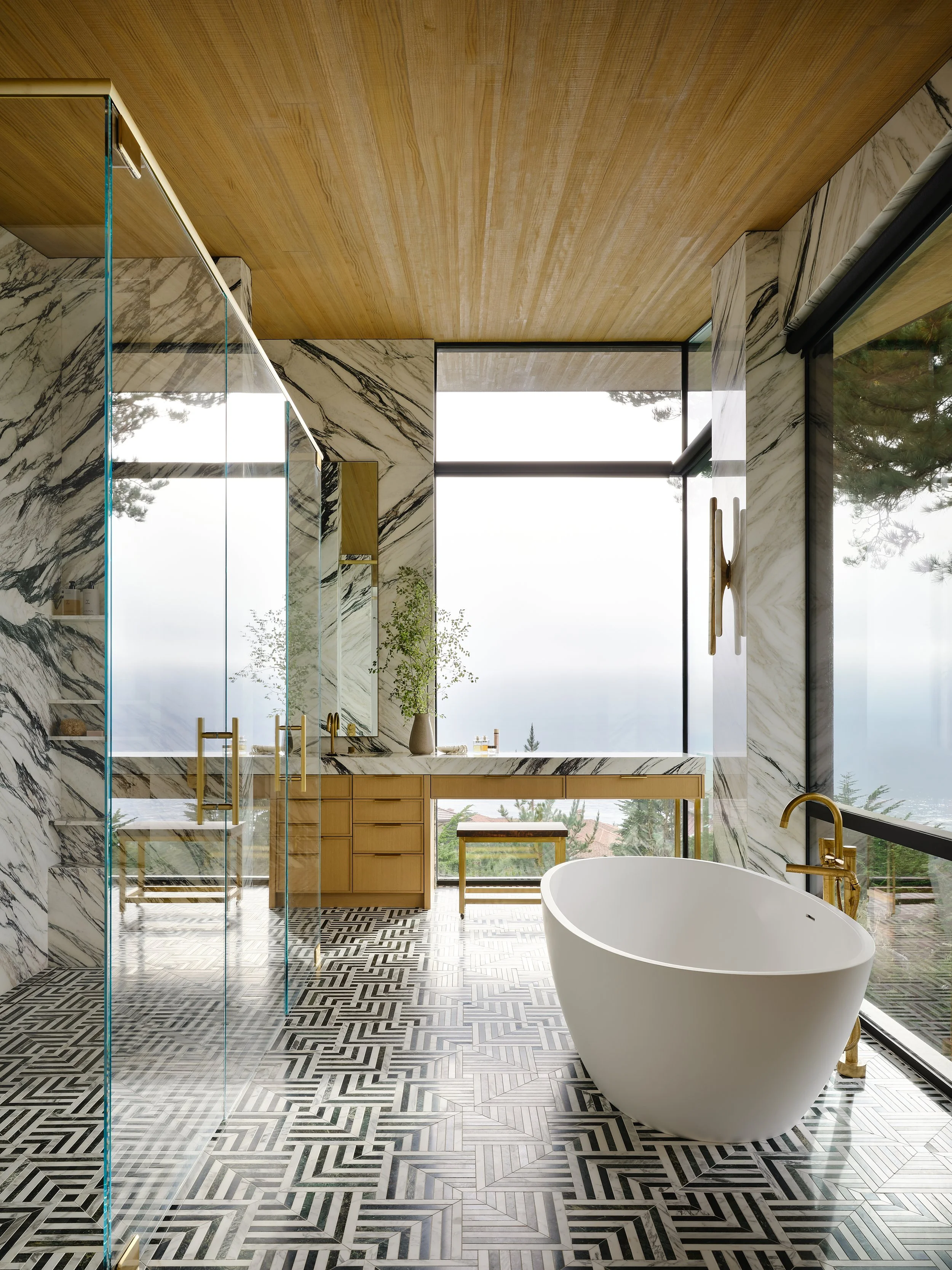 Modern bathroom with black and white patterned tile floor, a freestanding white bathtub with a gold faucet, large windows with a view of trees, a wooden ceiling, marble walls, a vanity with gold hardware, and a mirror.