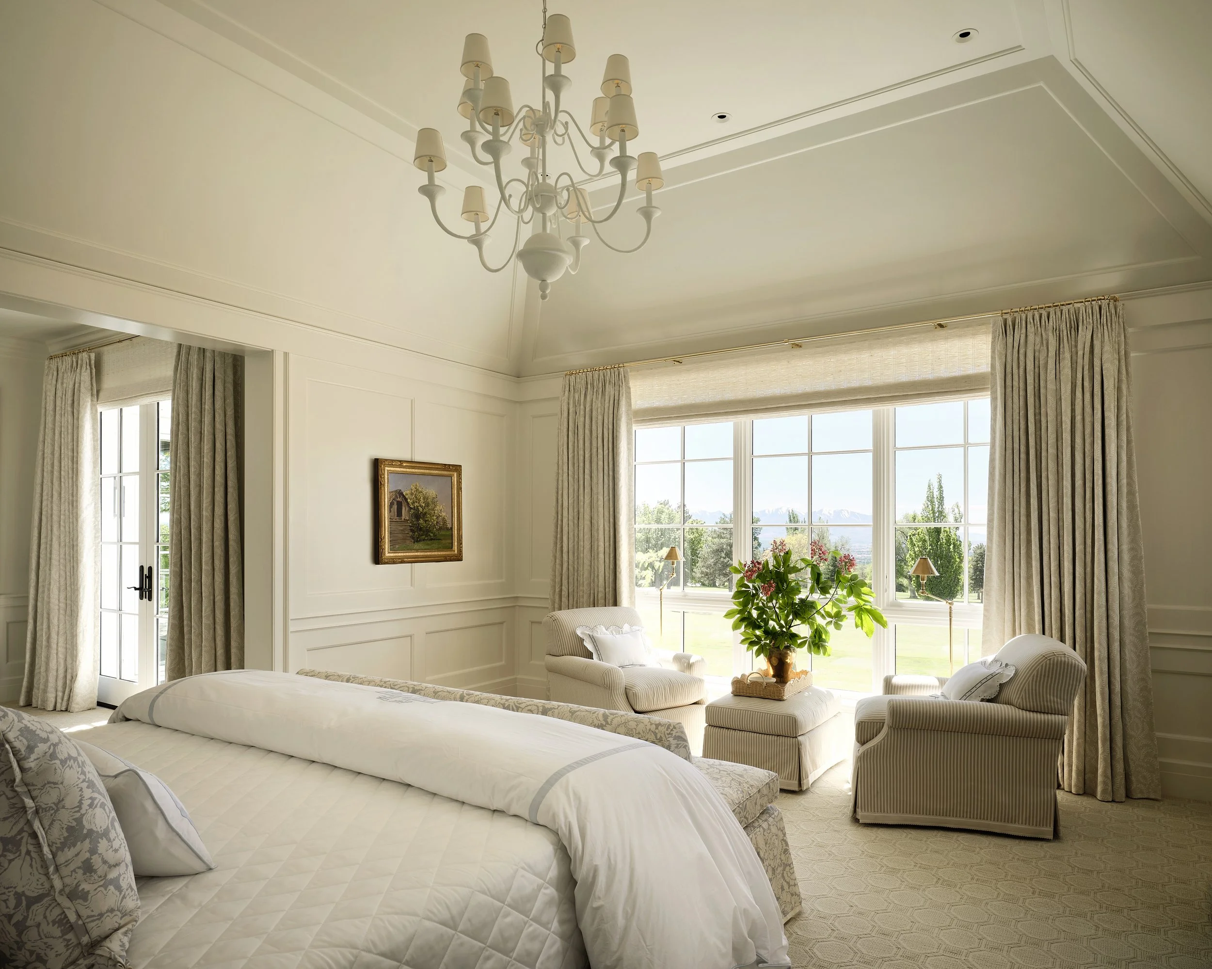 Bright bedroom with a large window, white curtains, and a floral arrangement on a small table between two armchairs. A bed with white bedding is in the foreground, and a chandelier hangs from the ceiling.
