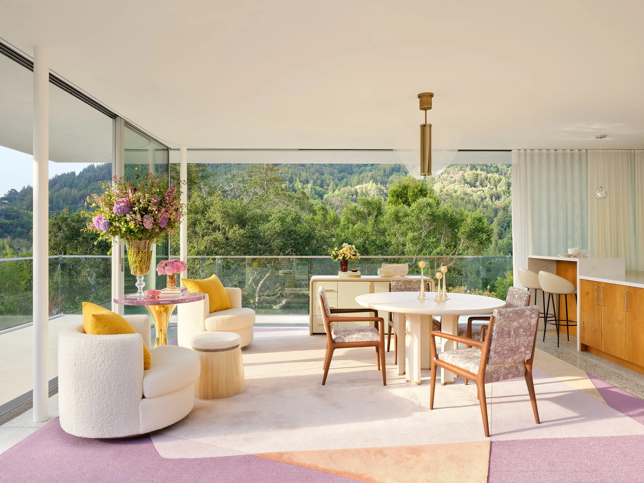 Living room with large windows showing a view of trees, featuring a round white table with yellow and pink flower arrangements, a white and yellow armchair with yellow pillows, and a dining area with four chairs around a white table. There's also a b