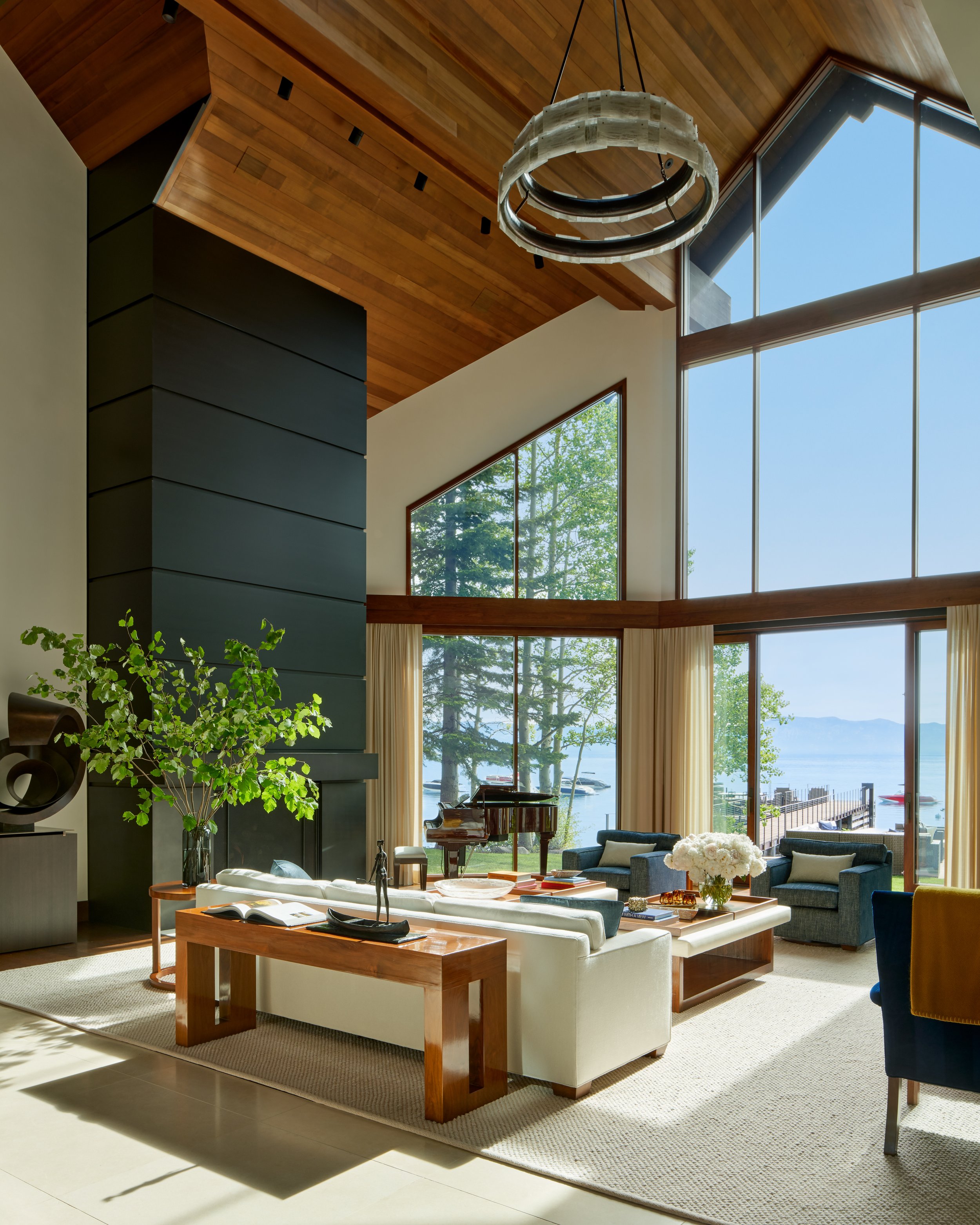 Living room with large floor-to-ceiling windows showing trees and water outside, modern furniture including a white sofa, a wooden coffee table with flowers, and a grand piano near the windows, high wooden ceiling with a circular chandelier.