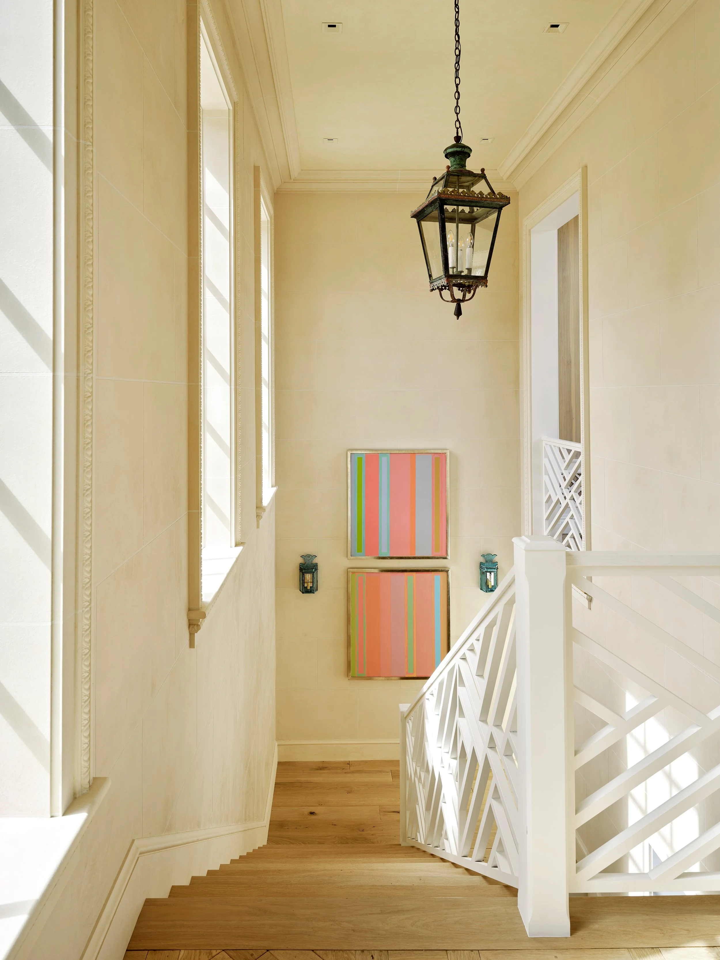 Bright staircase landing with beige walls, wooden flooring, a black lantern hanging from the ceiling, colorful abstract artwork on the wall, and white staircase railing and trim.