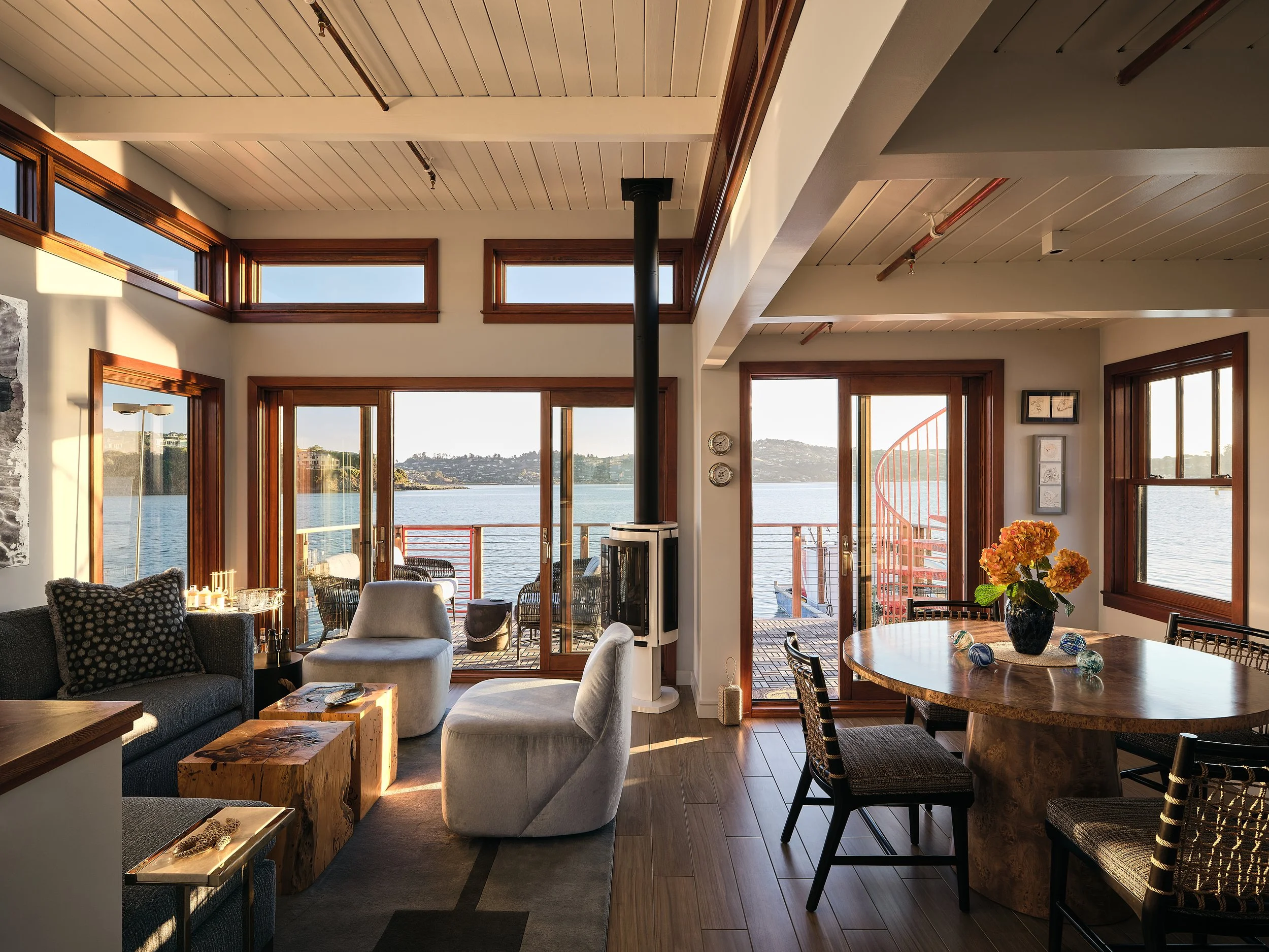 Living room with large windows and balcony overlooking a lake, with furniture including a sofa, armchairs, round wooden dining table with chairs, and a view of the outdoors.