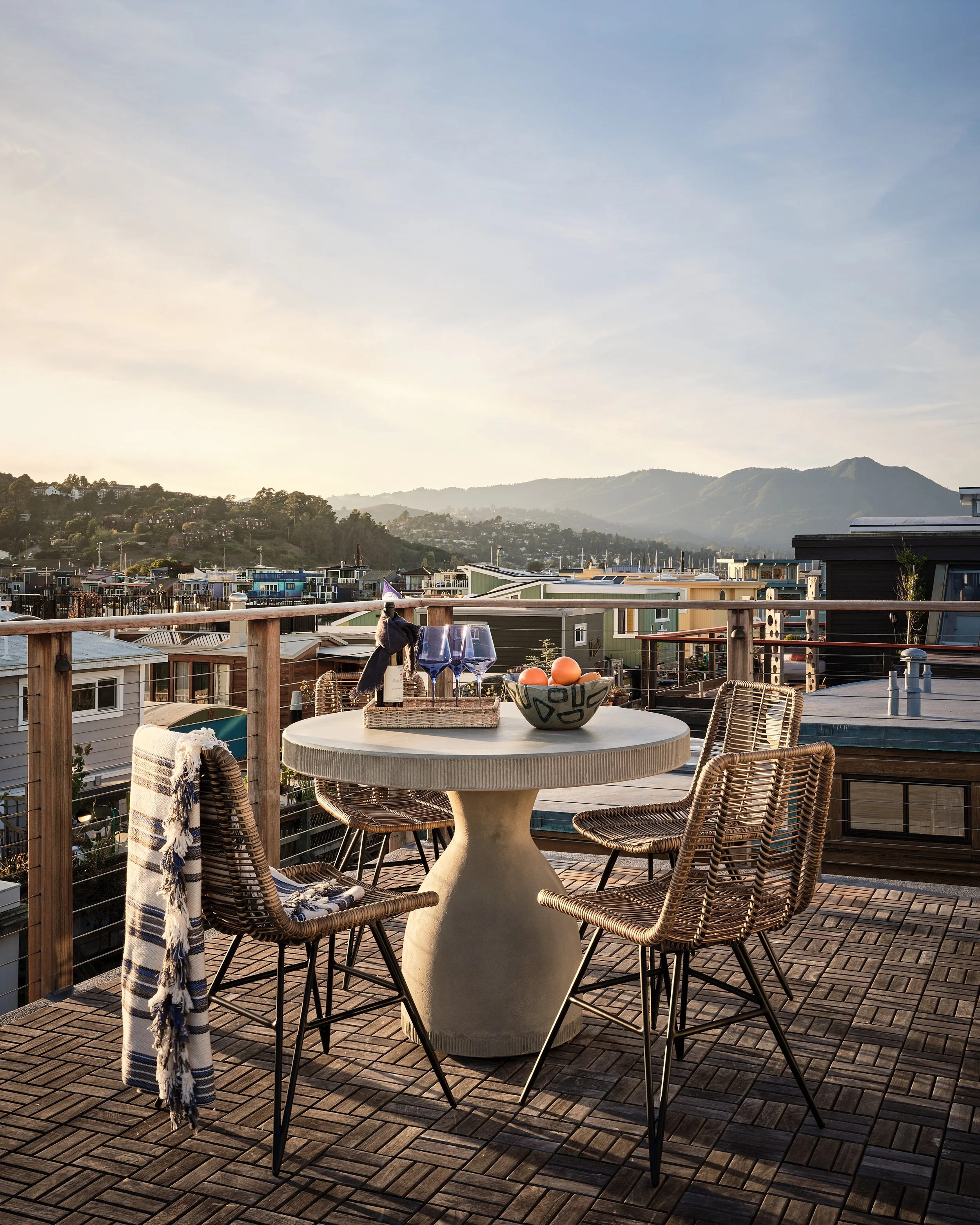 A rooftop patio with a round table and five wicker chairs, set with wine glasses, a bowl of oranges, and a basket of wine bottles, overlooking a view of a neighborhood and mountains under a partly cloudy sky at sunset.