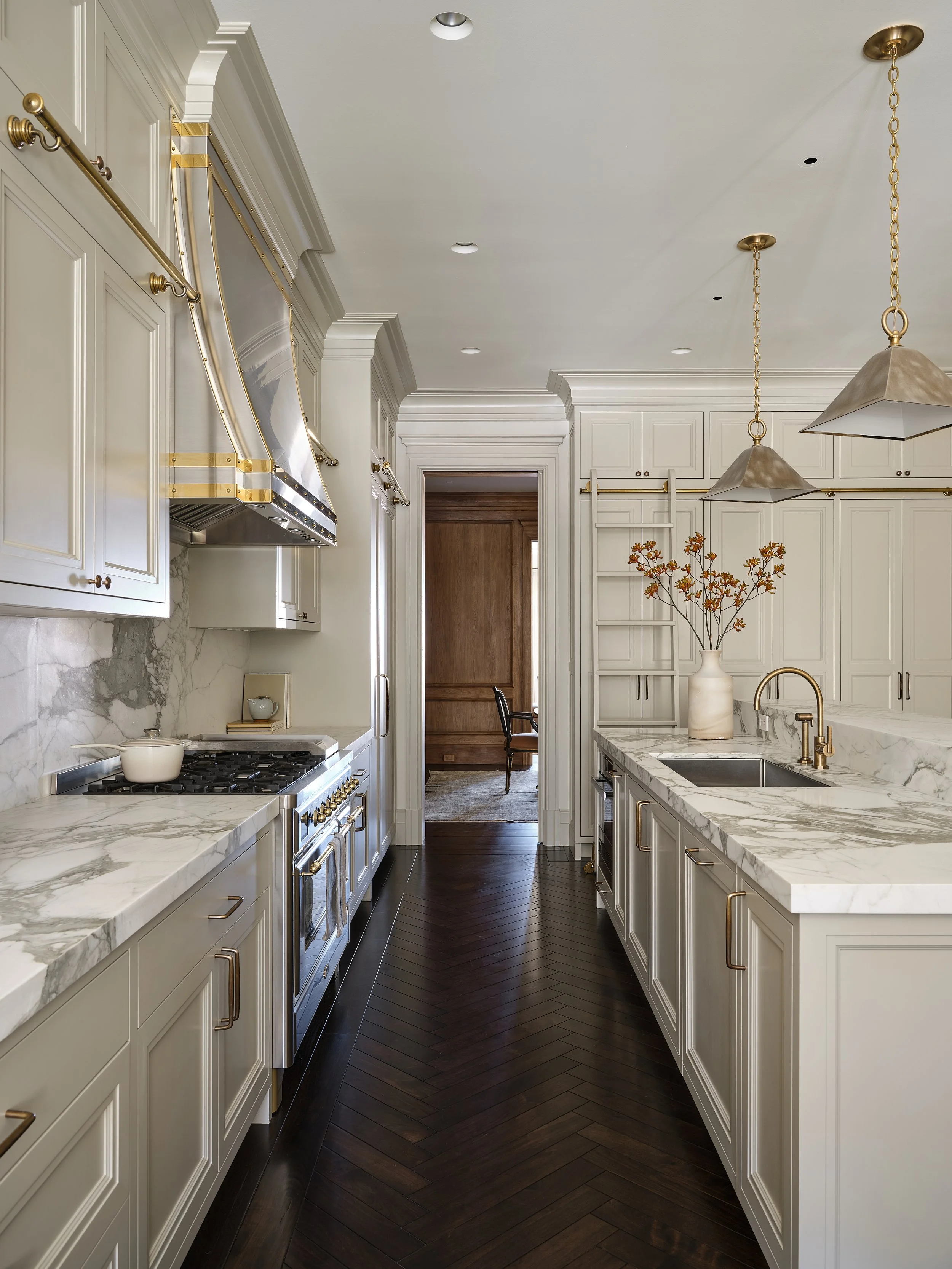 Modern kitchen with white cabinets, marble countertops, and gold hardware, featuring a stove, range hood, and pendant lights, with dark wood flooring and a doorway leading to another room.