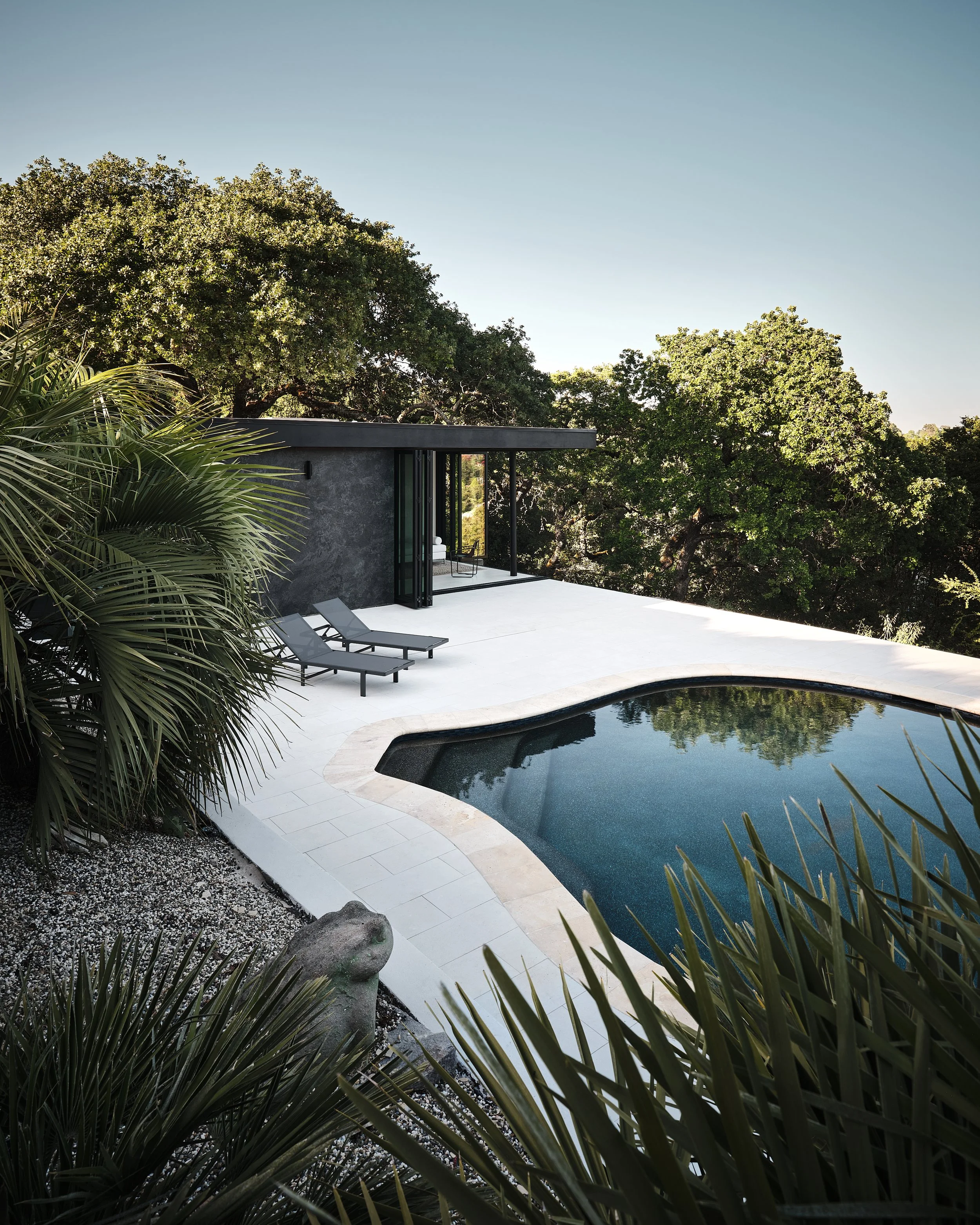 Modern backyard with a swimming pool, surrounded by lush trees, a small seating area, and a minimalist house with large glass doors.