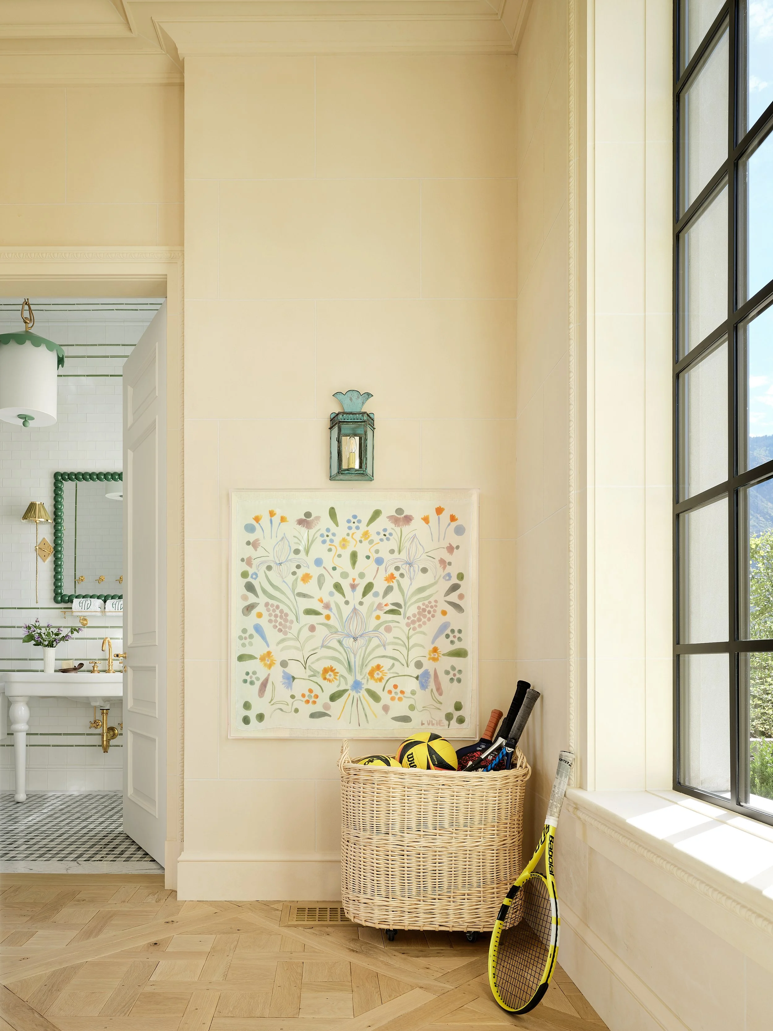 Interior corner of a room with a wicker basket filled with sports equipment, tennis racket, tennis balls, and baseballs, near a large window with a view outside, and a decorative wall art with colorful floral patterns.