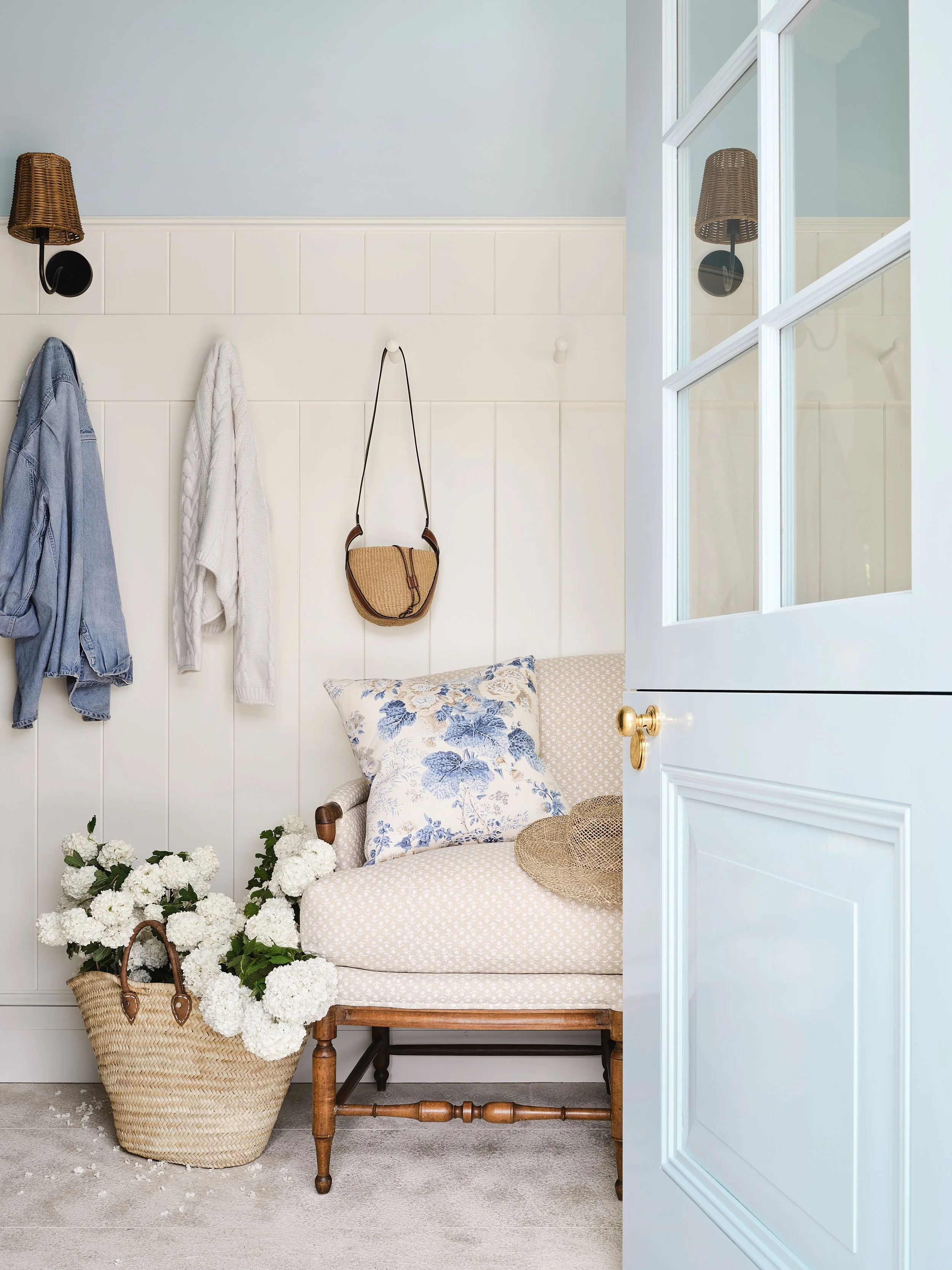 A cozy entryway with a white paneled wall, hanging clothing, a small purse, and a wicker hat on a vintage chair. A woven basket filled with white flowers is on the floor next to the chair, and a large mirror reflects the light blue walls and a wicker