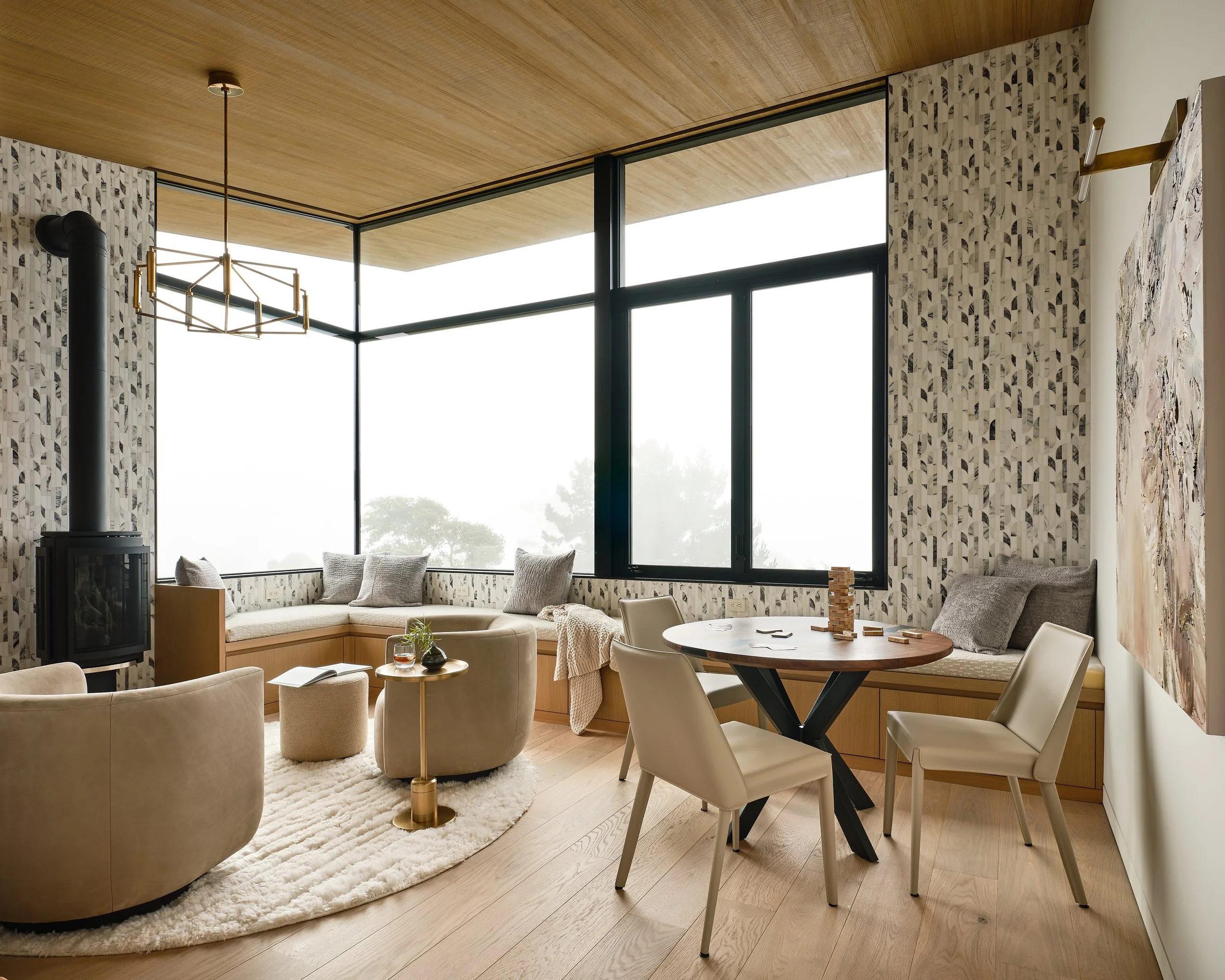Bright living room with large windows, modern furniture, a round table with chairs, a built-in window seat, patterned wallpaper, artwork, and a wood-burning stove. Interior design and Architecture. California architectural photographer