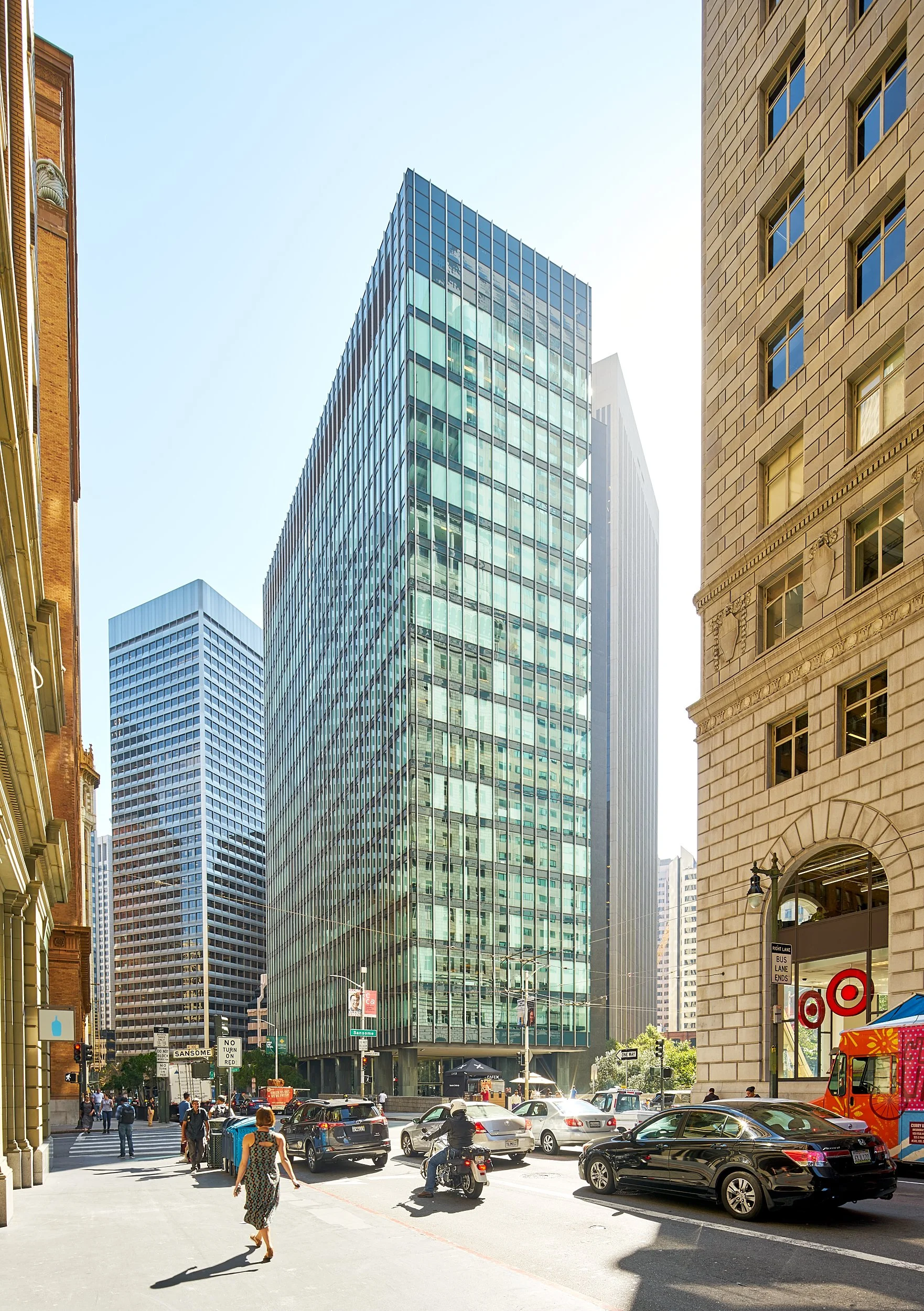 City street scene with tall glass office buildings, parked cars, a motorcycle, and pedestrians walking on the sidewalk.
