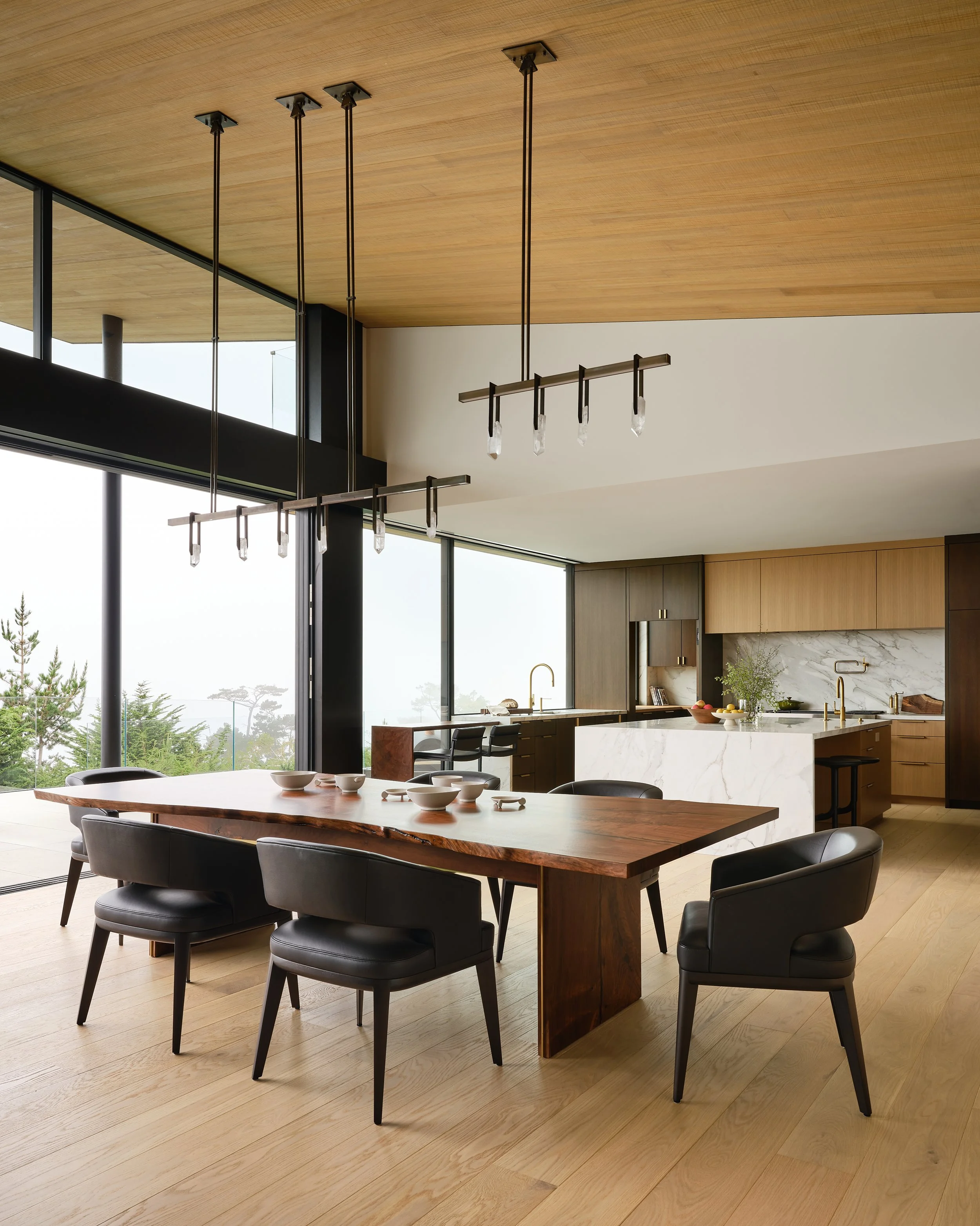 Modern open-concept kitchen and dining area with a large wooden dining table, black chairs, pendant lighting, marble kitchen island, wooden cabinets, and large windows showing trees outside.