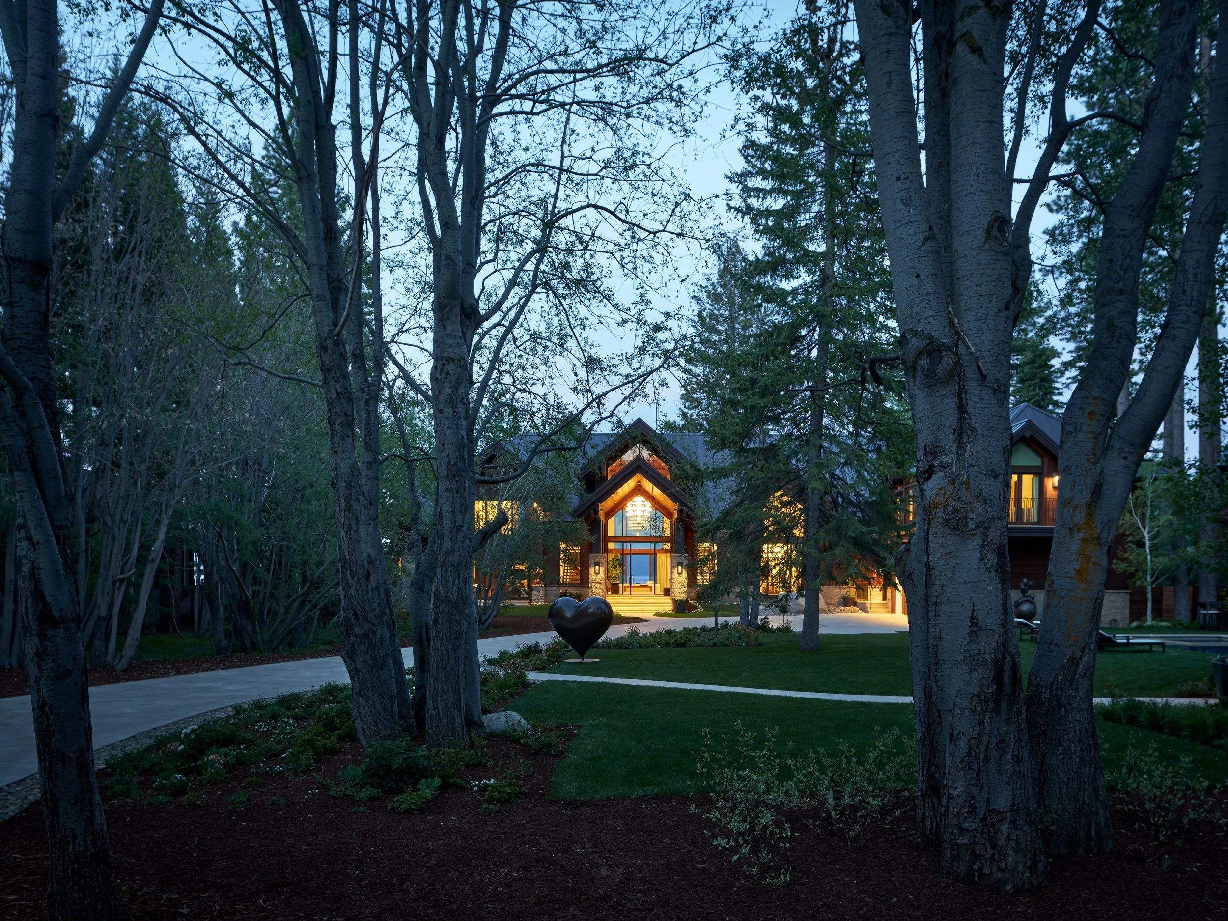 A house illuminated at dusk, surrounded by trees and a landscaped yard with a paved walkway.