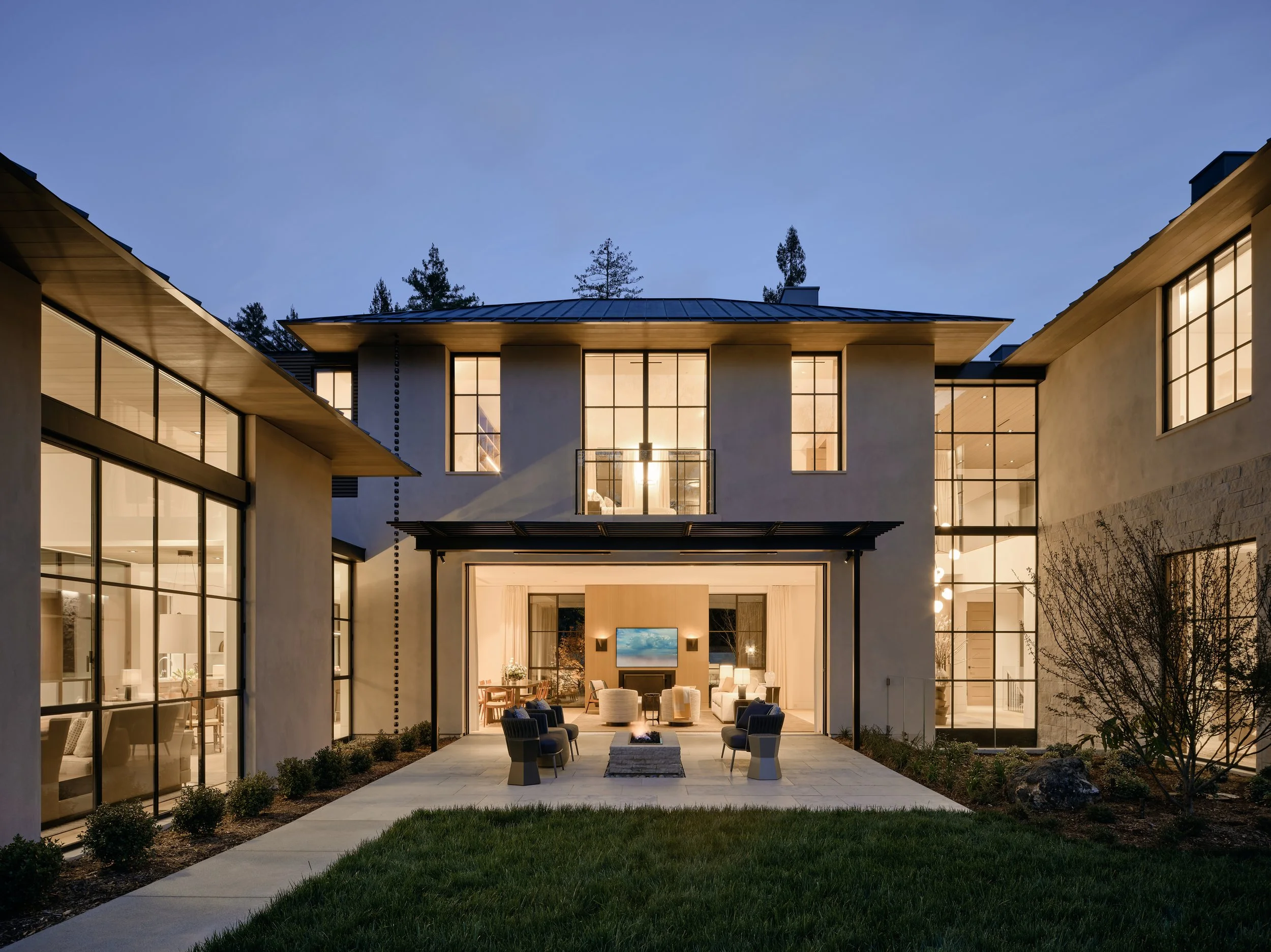 Modern multi-story house with large glass windows, lit interior, outdoor patio with seating area, grass lawn, and trees in the background at dusk. Interior design and Architecture. California architectural photographer