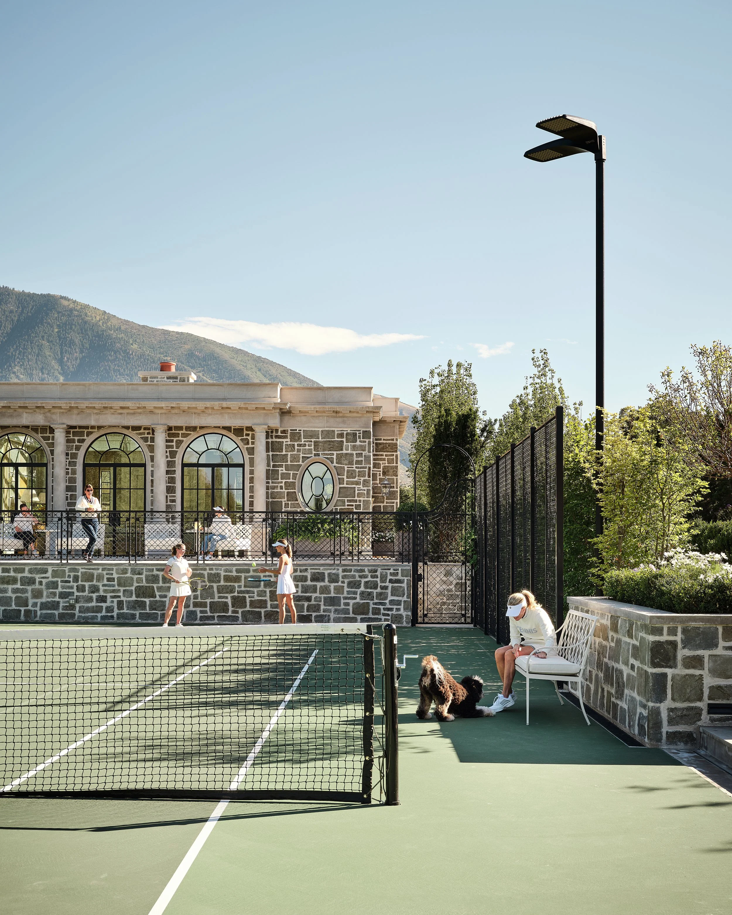 People playing tennis on an outdoor court with a woman sitting on a bench with a dog beside her, and a building with arched windows in the background.