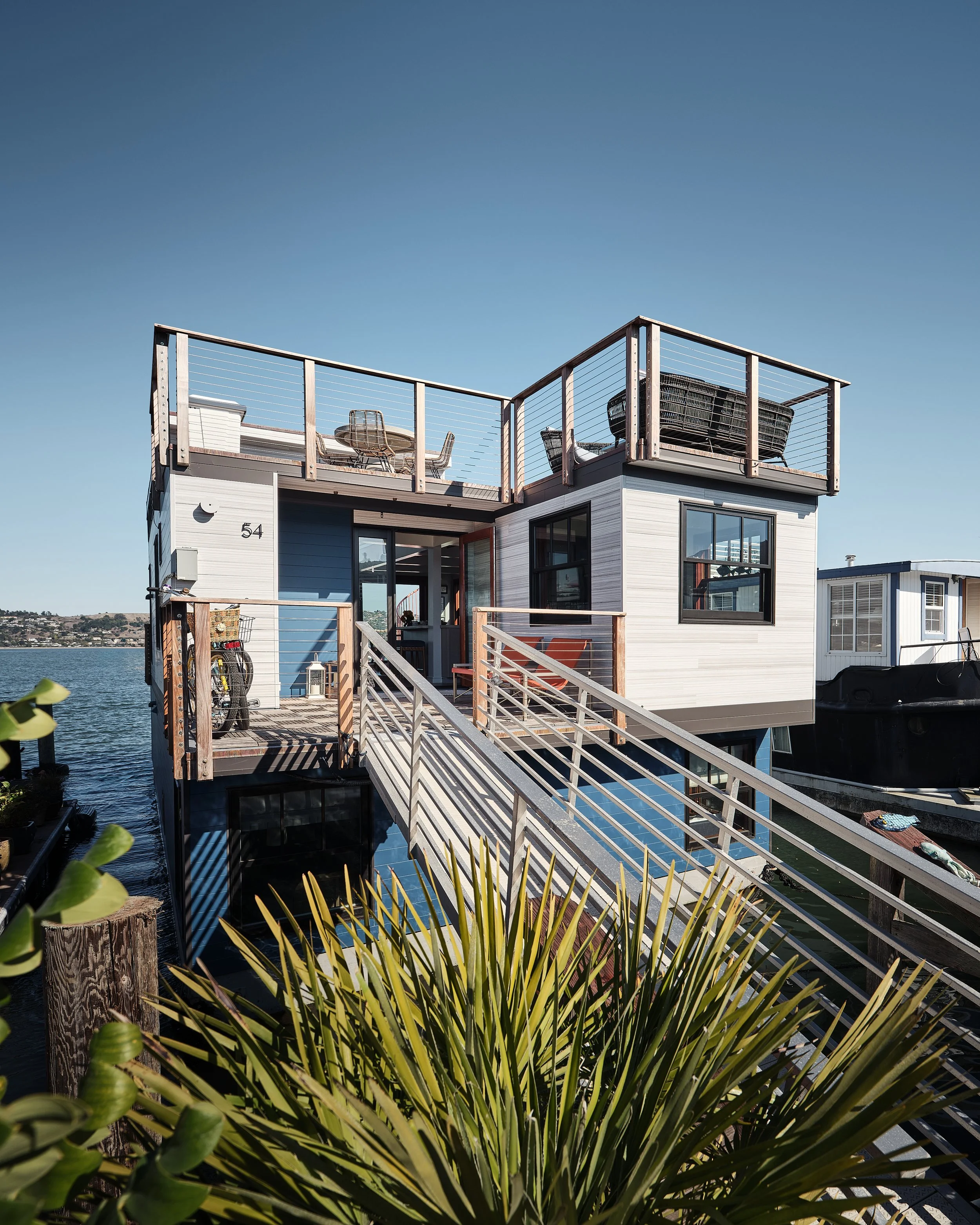 A modern floating house with a wooden staircase leading to a deck overlooking a body of water. The house has large windows, a rooftop patio with chairs, and a blue and white exterior.